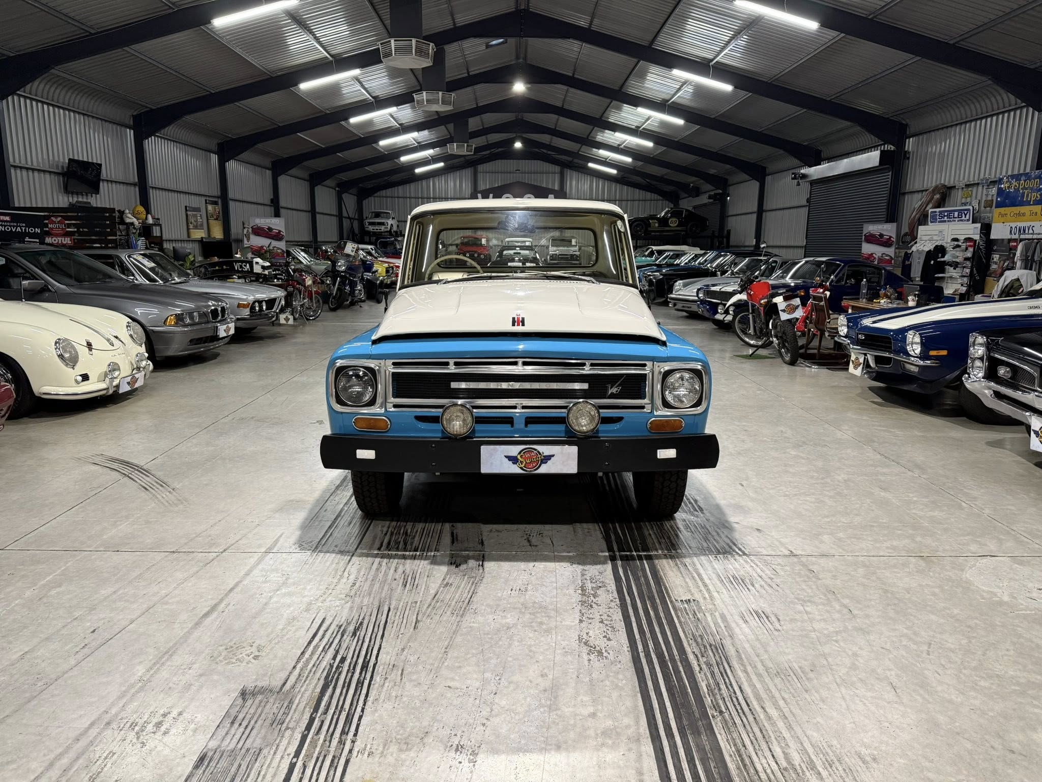 Front view of a blue-and-white vintage pickup in a large showroom surrounded by numerous classic cars and motorcycles on display.