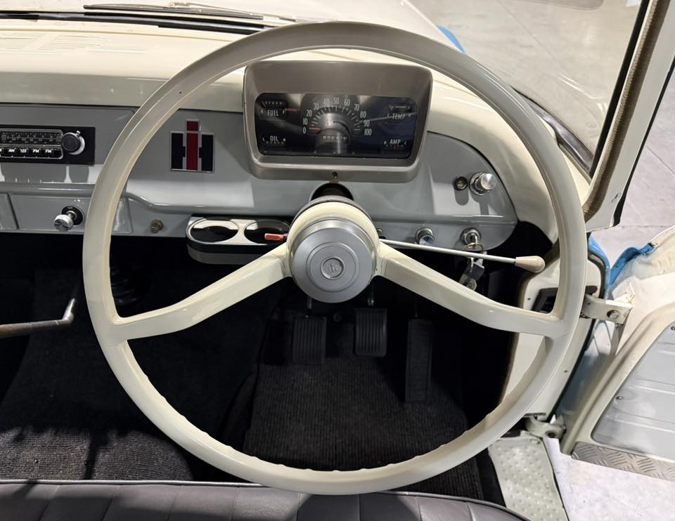 Vintage car interior showing a large cream steering wheel, grey dashboard, and analog gauges behind it.