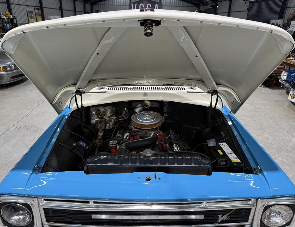 Open-hood view of a blue vintage car showing its engine bay with a round air cleaner on top of the engine.