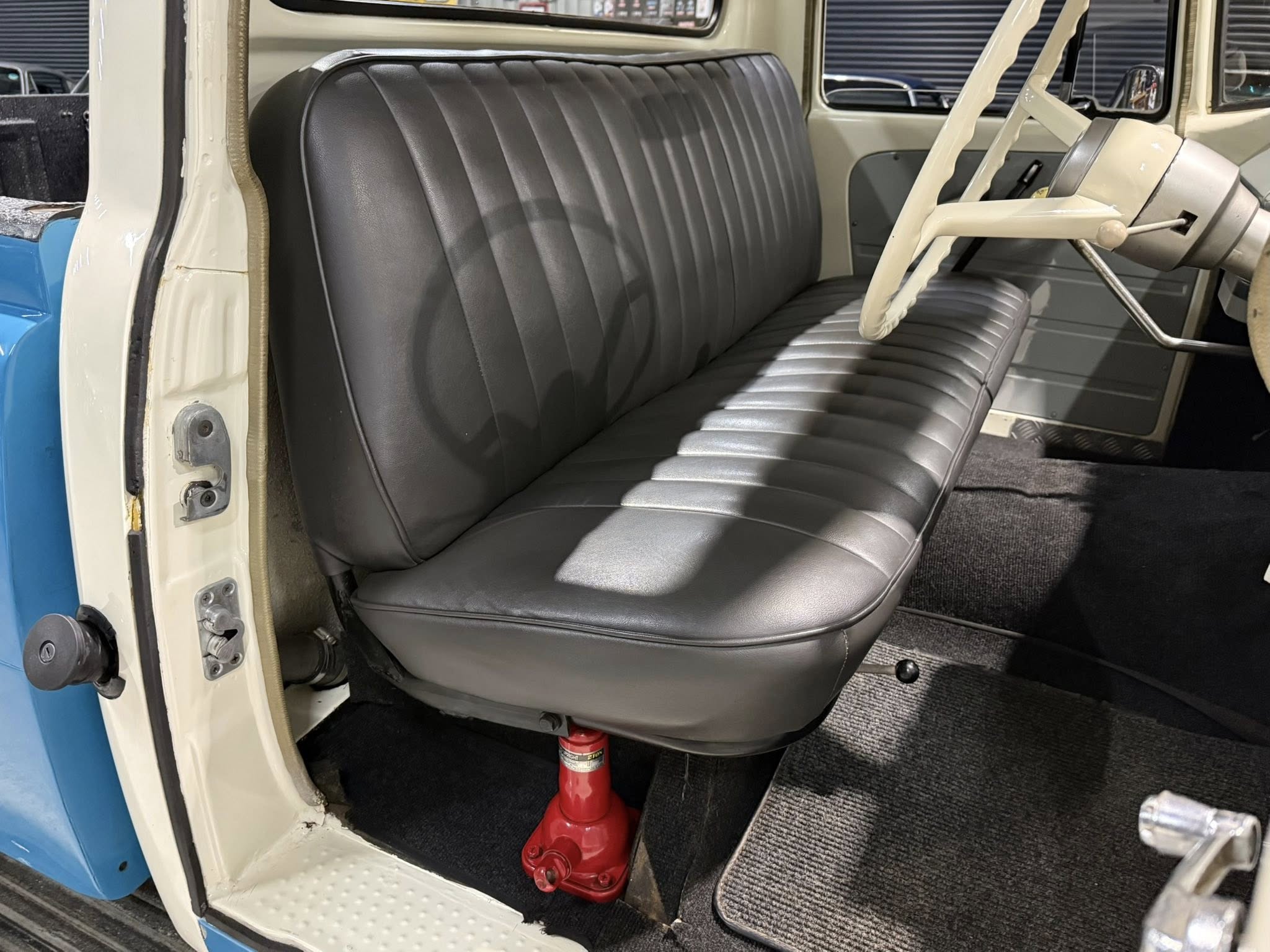 Vintage truck cab interior with gray bench seat, large steering wheel, and a red hydraulic jack under the driver’s seat outside the door frame.