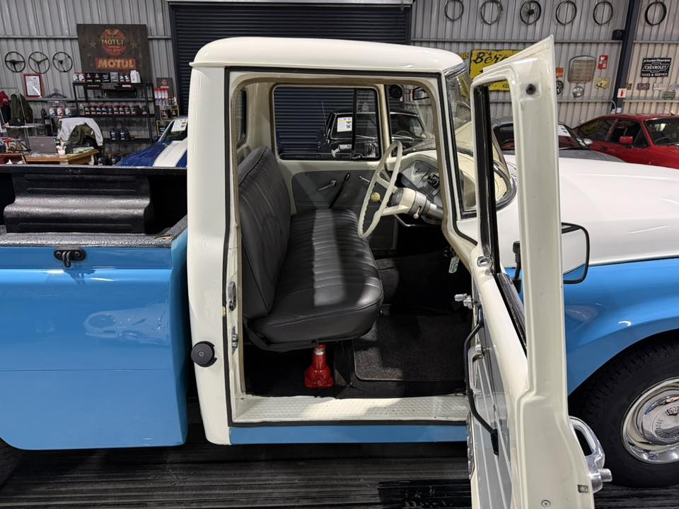 Vintage blue and white pickup truck with the driver's door open, showing a gray bench seat and vintage steering wheel inside a workshop area.