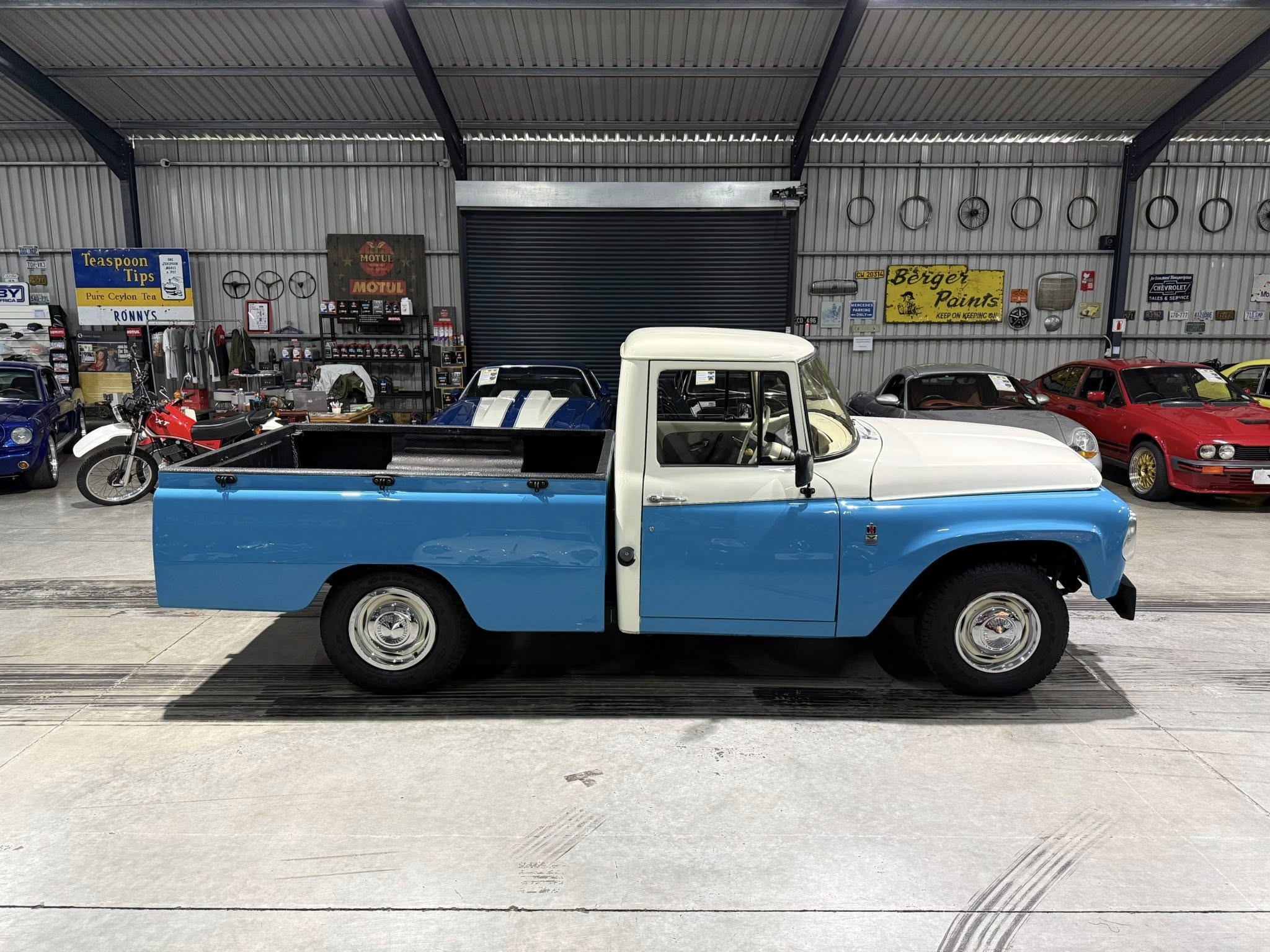 Blue and white vintage pickup truck in a garage showroom, with retro signs, bikes hanging on the wall, and other cars in the background.