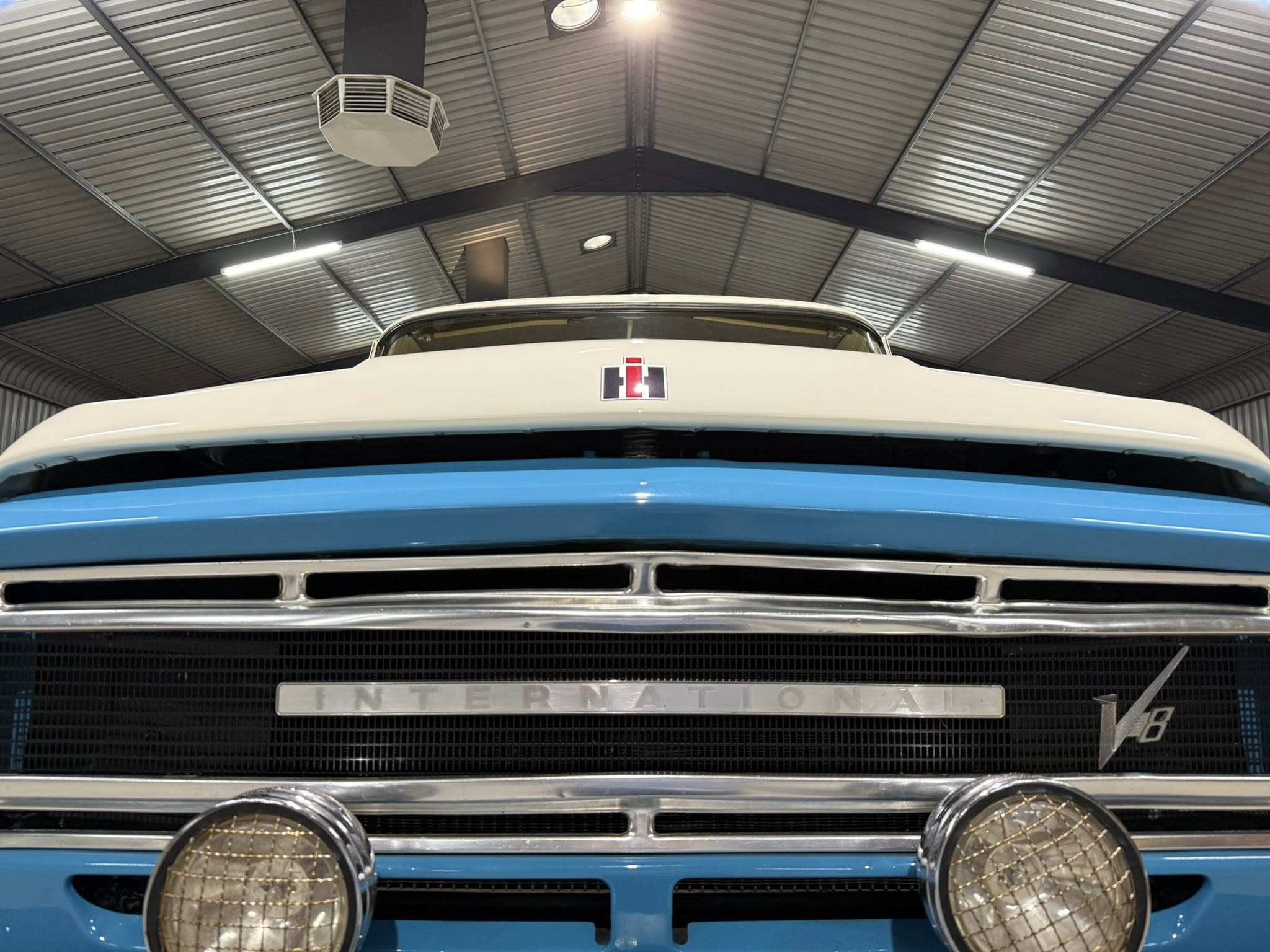 Front view of a vintage white and blue International bus inside a metal warehouse, showing the iconic grille and round headlights with protective grills in the foreground.