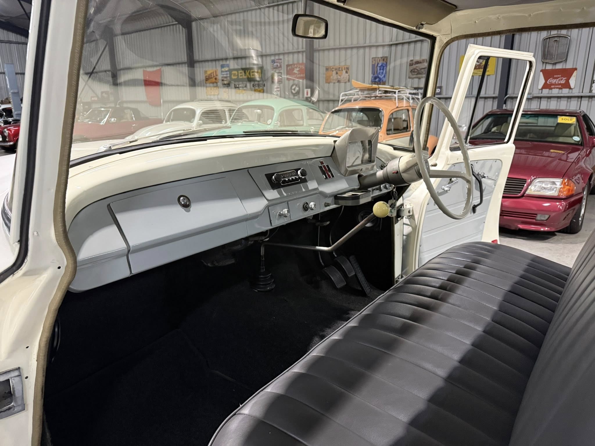 Interior of a vintage car with the driver's door open, showing a gray dashboard, steering wheel, and bench seat; other colorful classic cars visible in the background showroom.
