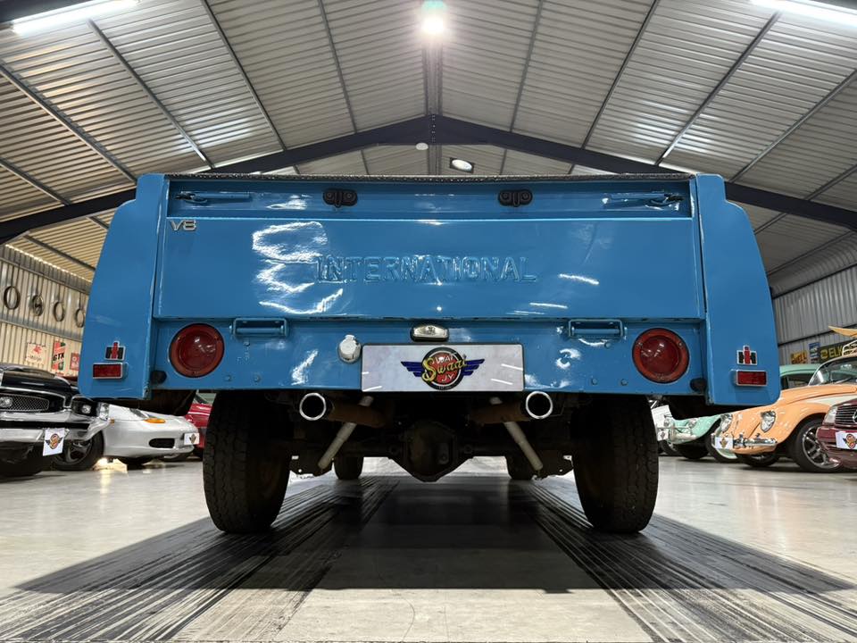 Blue vintage International pickup viewed from the rear in an indoor showroom, with other classic cars lining the sides.