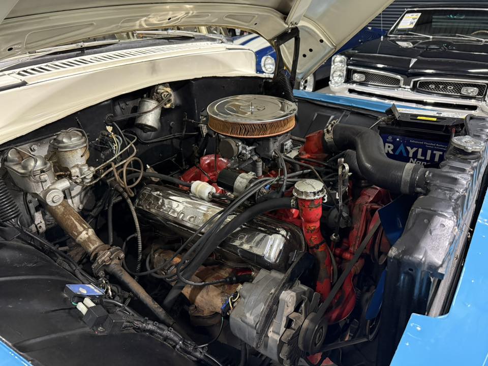 Classic car engine bay with a chrome air filter and red intake manifold; blue exterior visible at edge.
