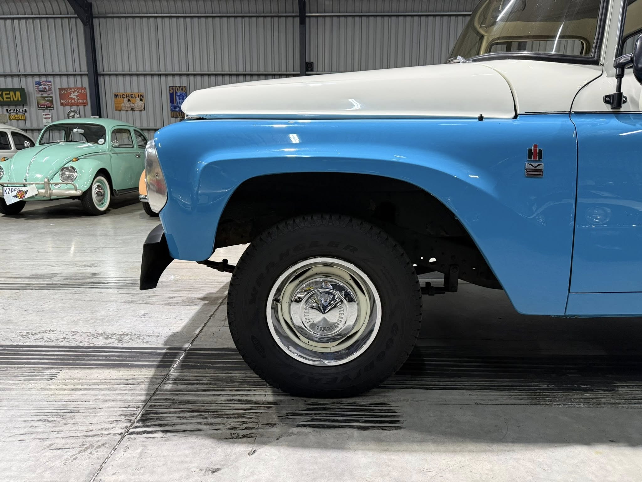 Close-up of a blue vintage truck fender and front tire inside a garage with classic cars in the background.
