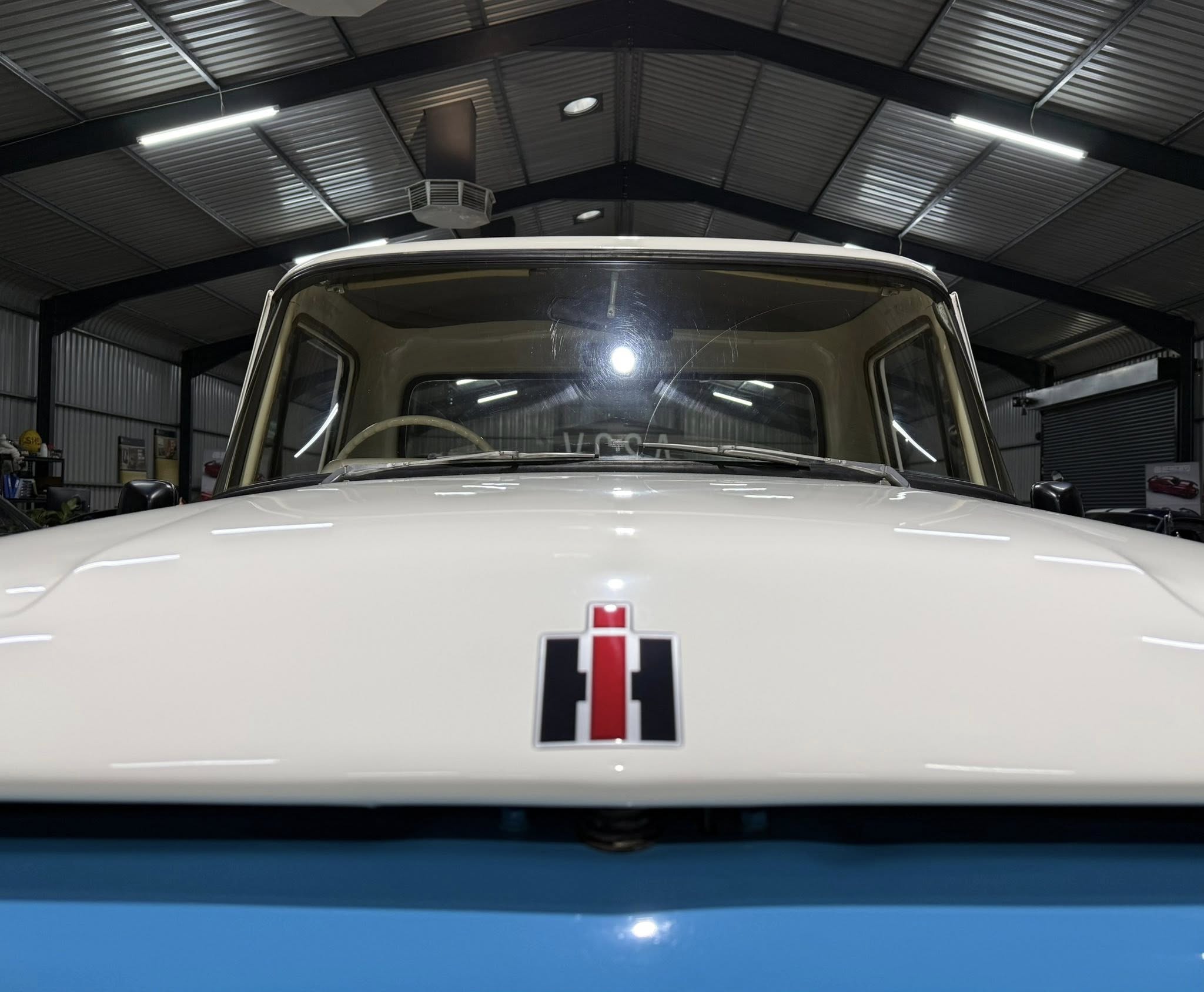 Front of a vintage bus in a workshop, hood emblem with red vertical stripe visible under overhead lighting.