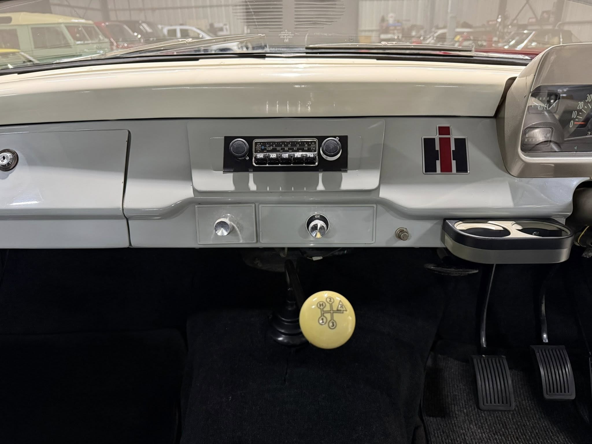 Vintage car dashboard viewed from the driver's seat, with a retro radio, heater controls, a yellow gear-shift knob, and three pedals visible below.