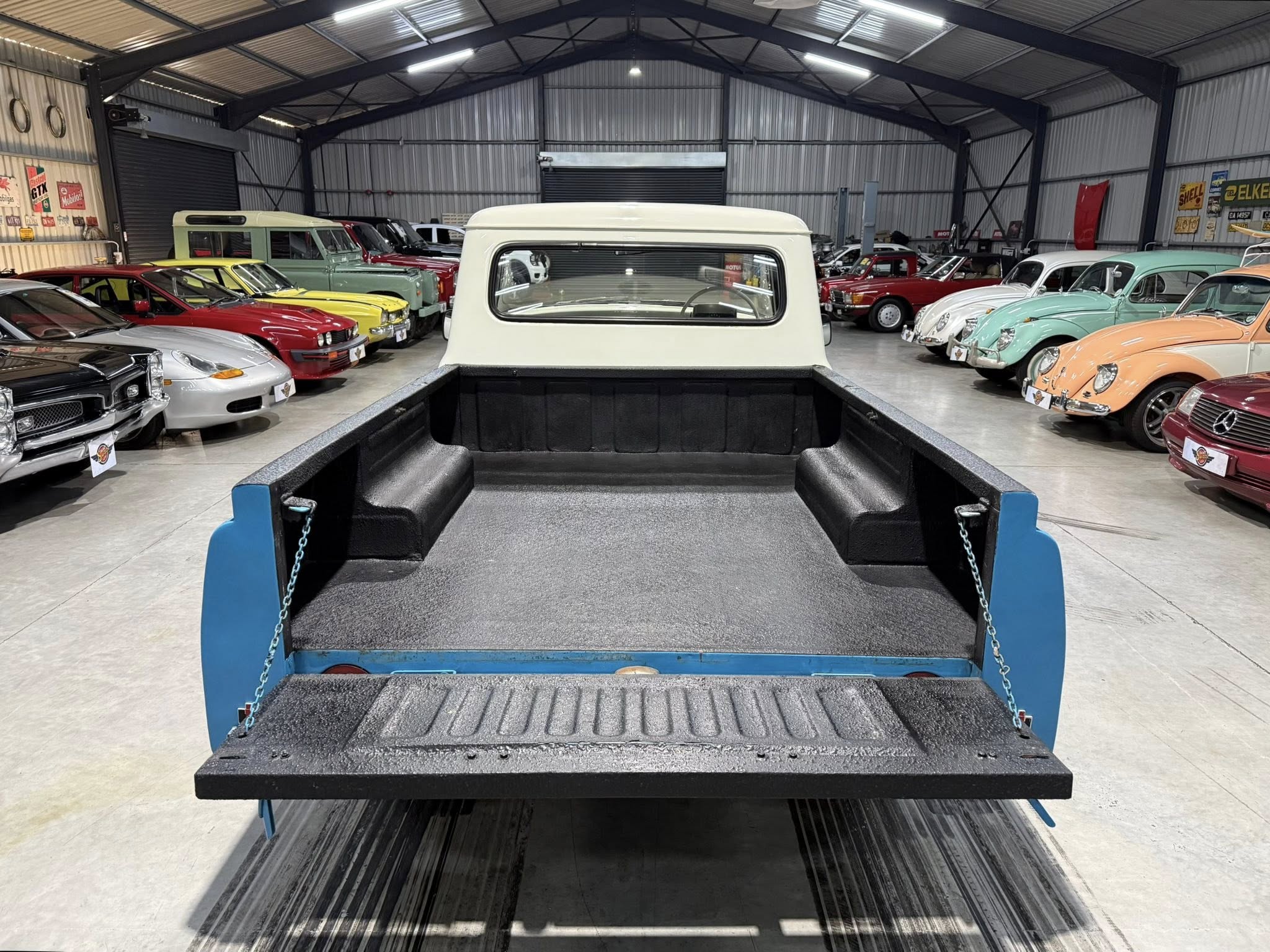 View from the bed of a blue pickup truck looking over a row of vintage cars inside a showroom warehouse.