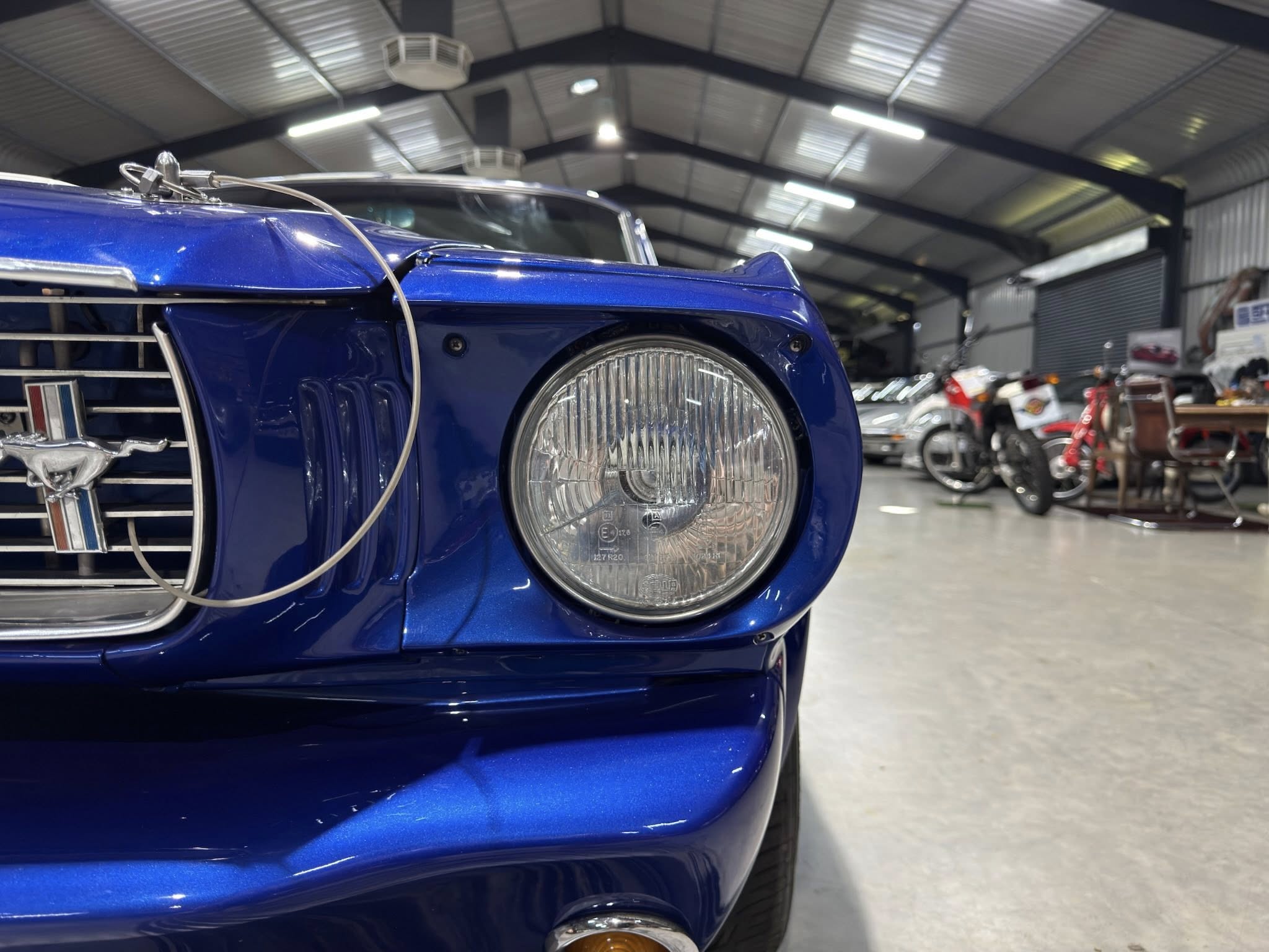 Close-up of a blue vintage car front left, showing the headlight and chrome grill in a showroom with motorcycles in the background.