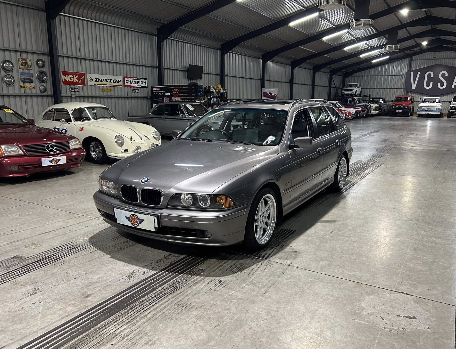 Indoor showroom of vintage cars, with a silver BMW wagon in the foreground and classic cars lined along the sides under bright lights, banners on the wall.