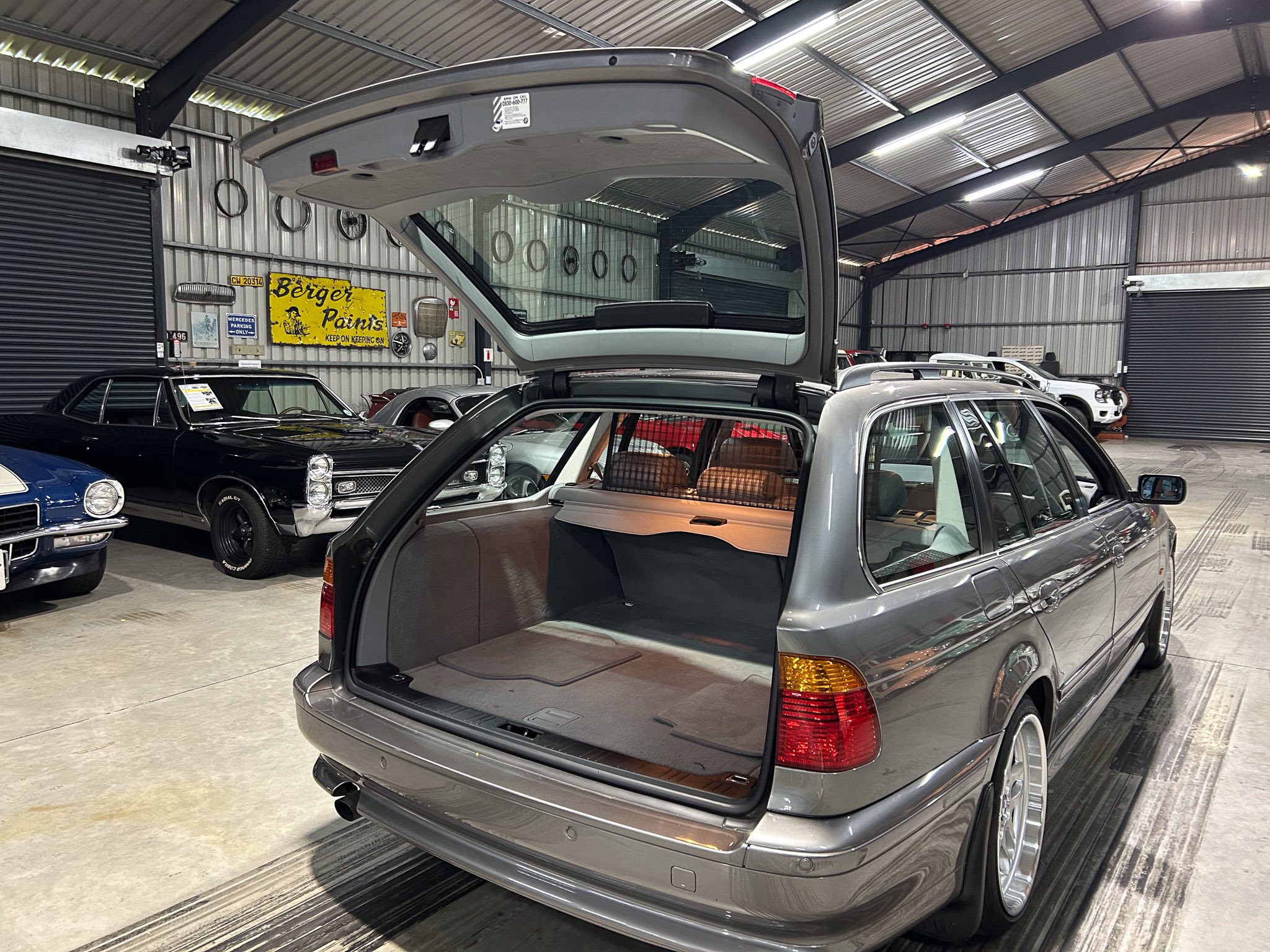 Gray SUV with its rear hatch open, exposing the cargo area inside a car showroom or workshop with other classic cars nearby and industrial walls with hanging tires in the background.