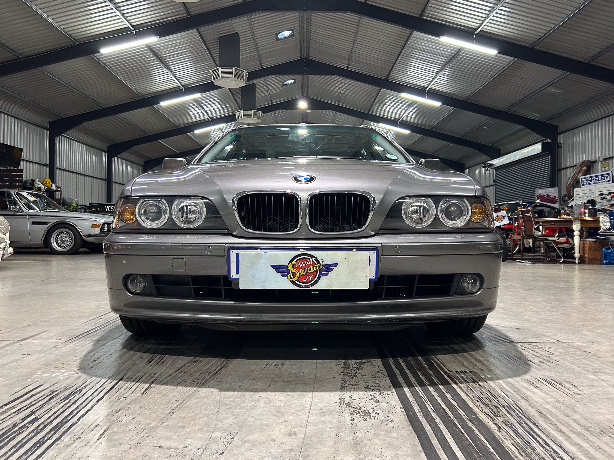 Front view of a silver BMW inside a car showroom with other vehicles and workshop equipment in the background.