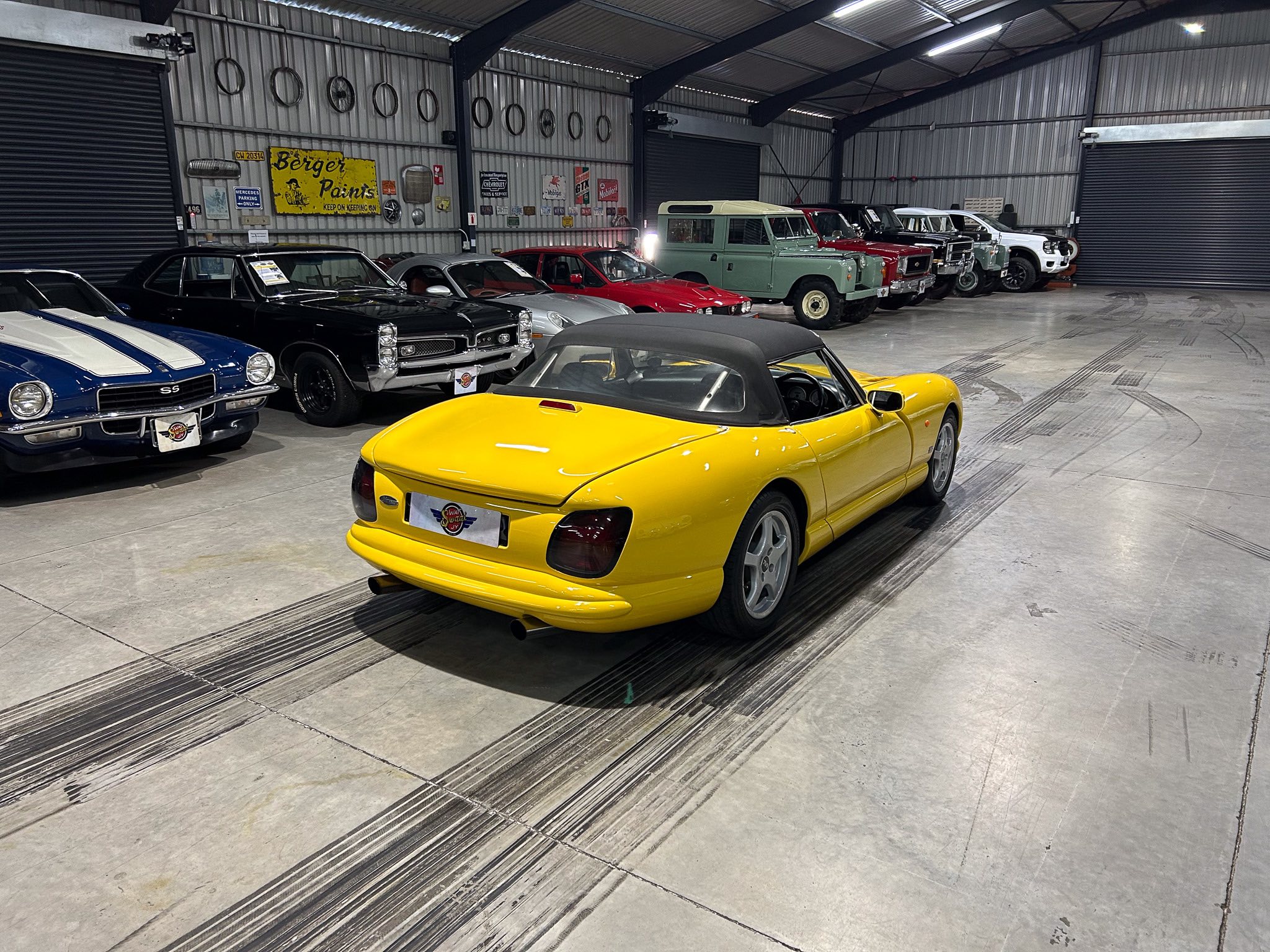 Indoor car showroom with a bright yellow convertible in the foreground and a row of vintage cars along the right wall of a metal warehouse.