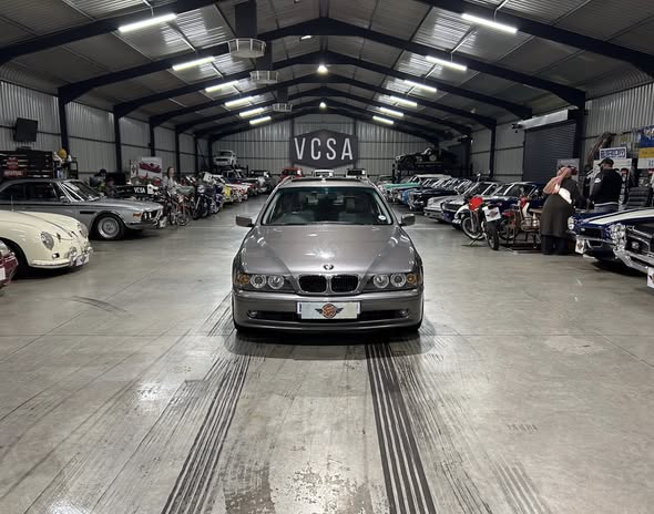 Front view of a silver BMW sedan inside a large indoor car showroom, with classic cars lined on both sides and a VCSA sign visible in the back.