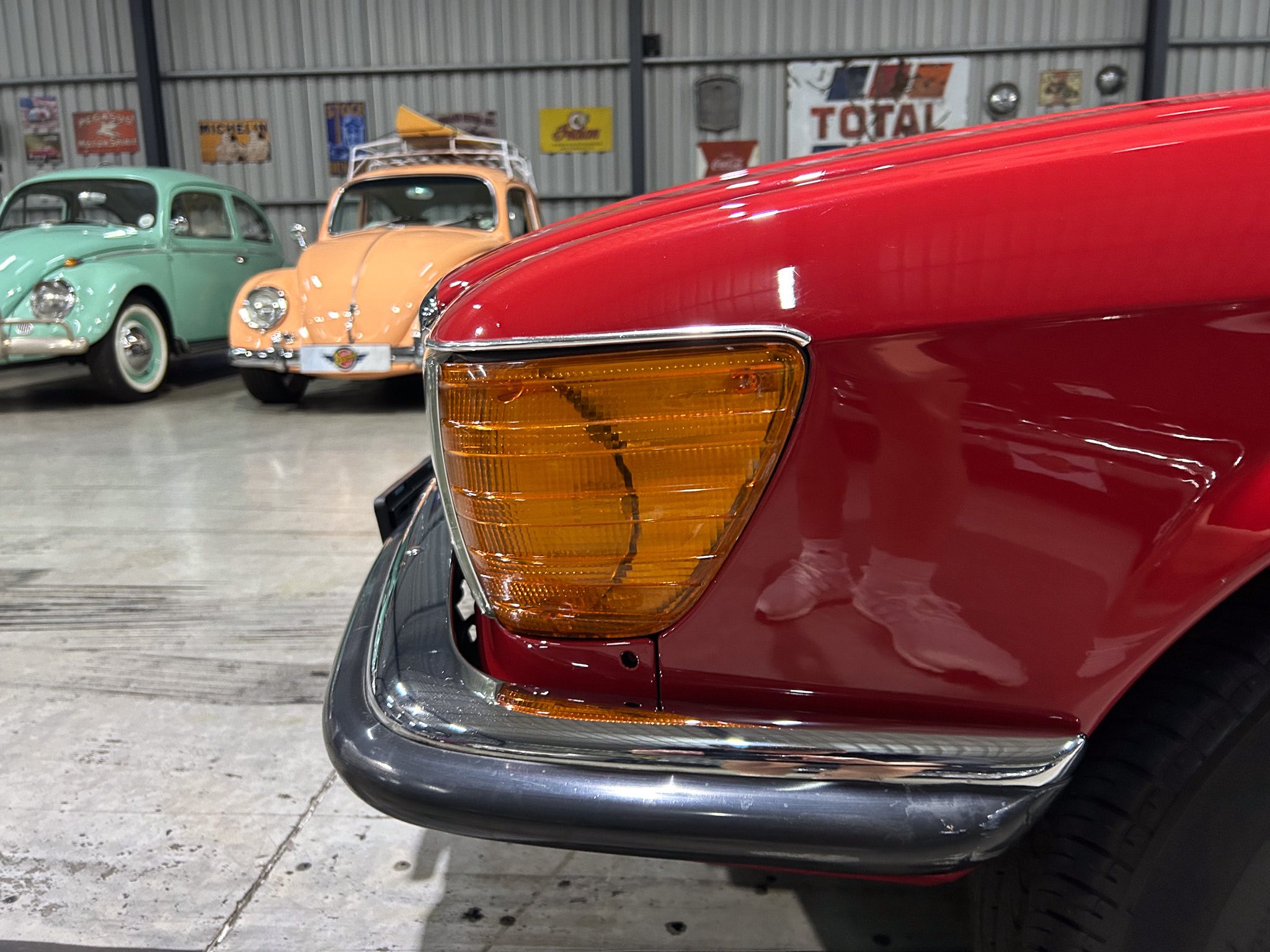 Close-up of a red classic car's front corner showing the amber turn signal and chrome bumper in a vintage showroom with other old cars in the background.