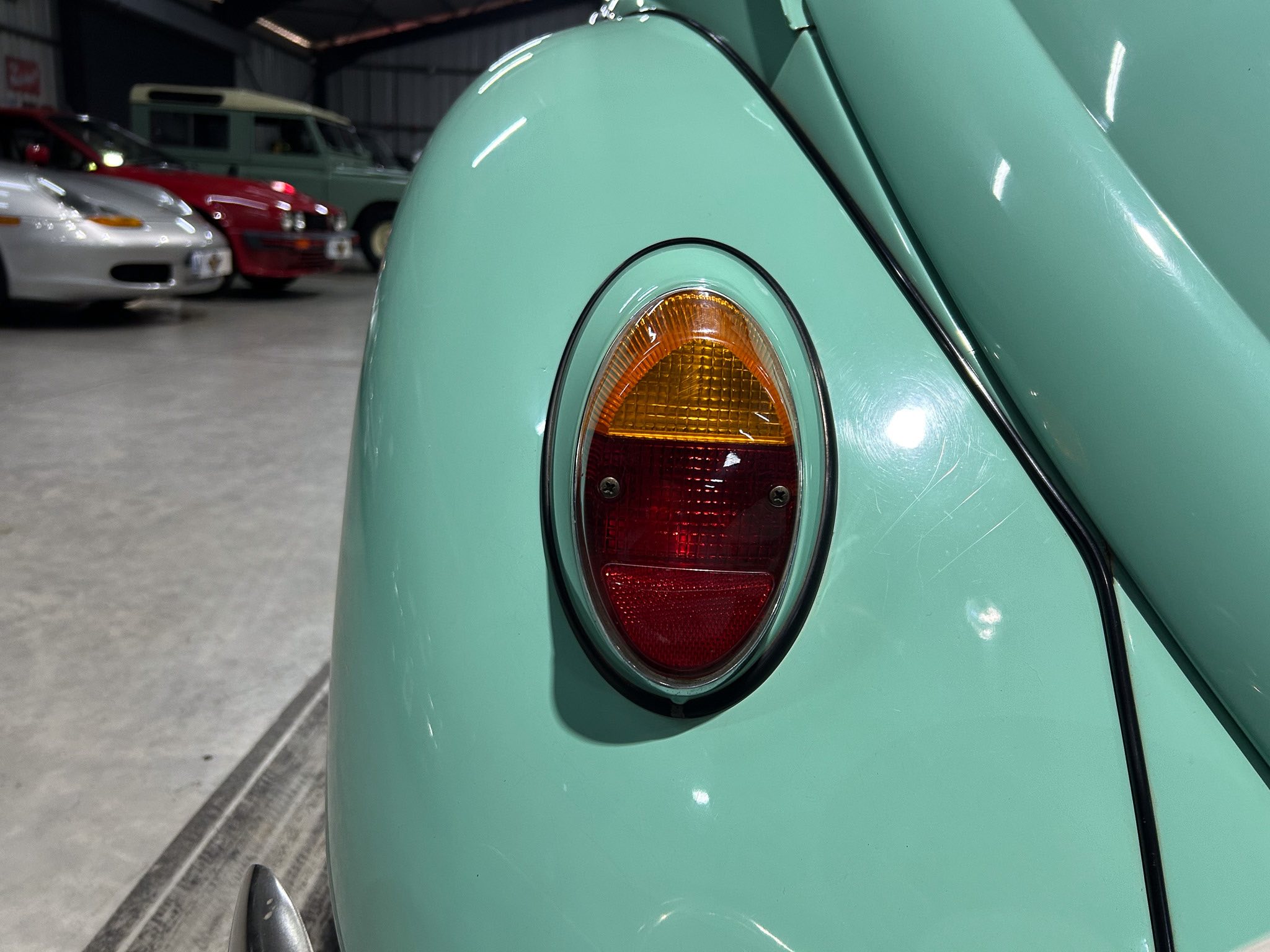 Close-up of a mint-green vintage car's tail light and curved body panel in a garage, with other cars blurred in the background.