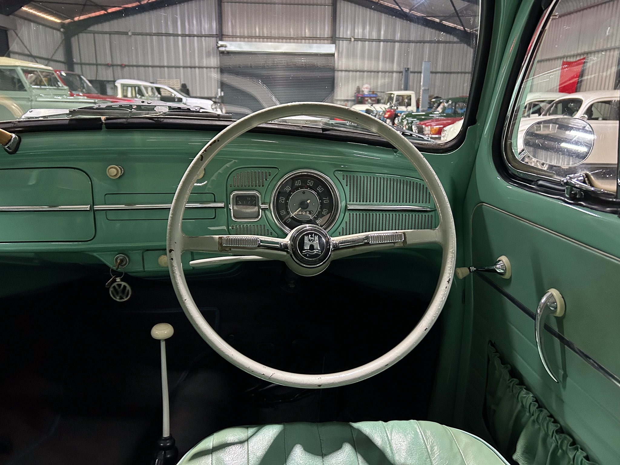 Green vintage car interior seen from the driver’s seat, with a white steering wheel and classic dashboard gauges inside a showroom garage.