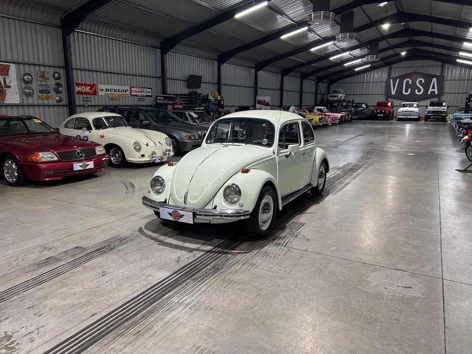 White vintage Volkswagen Beetle parked in a spacious indoor showroom surrounded by other classic cars in a museum-like setting.