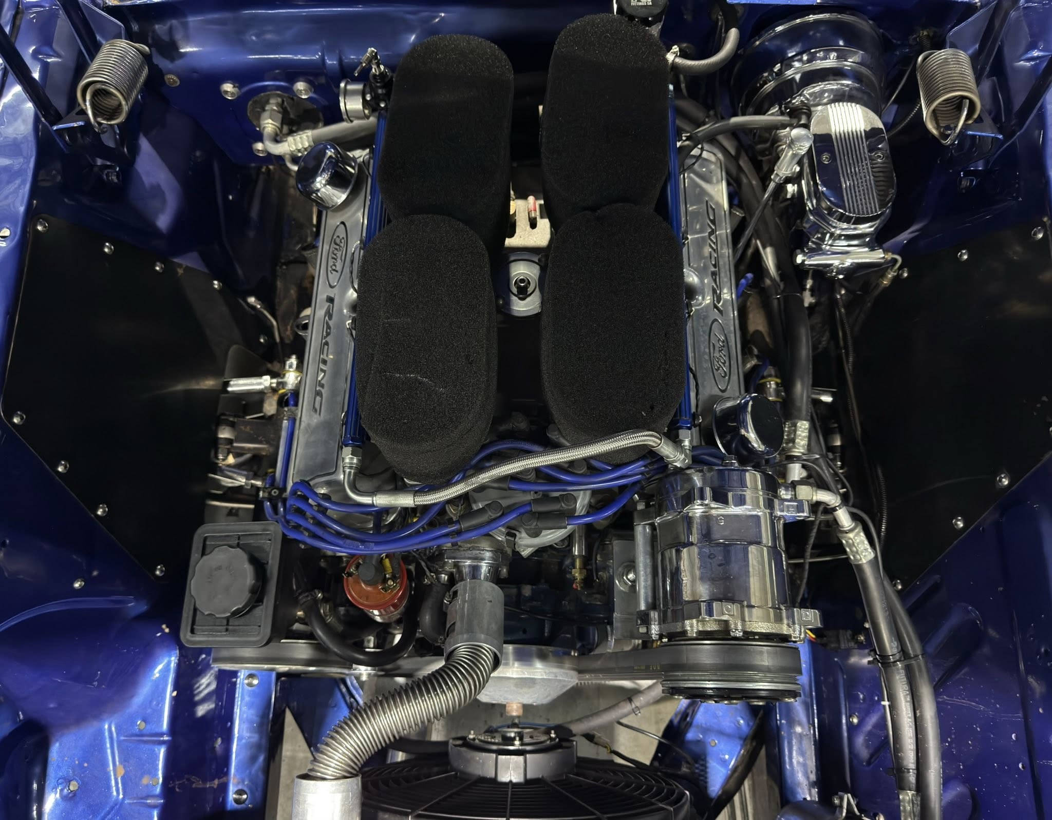 Blue race car engine bay with twin black foam air intake cushions at center, chrome engine parts, and blue braided hoses surrounding them