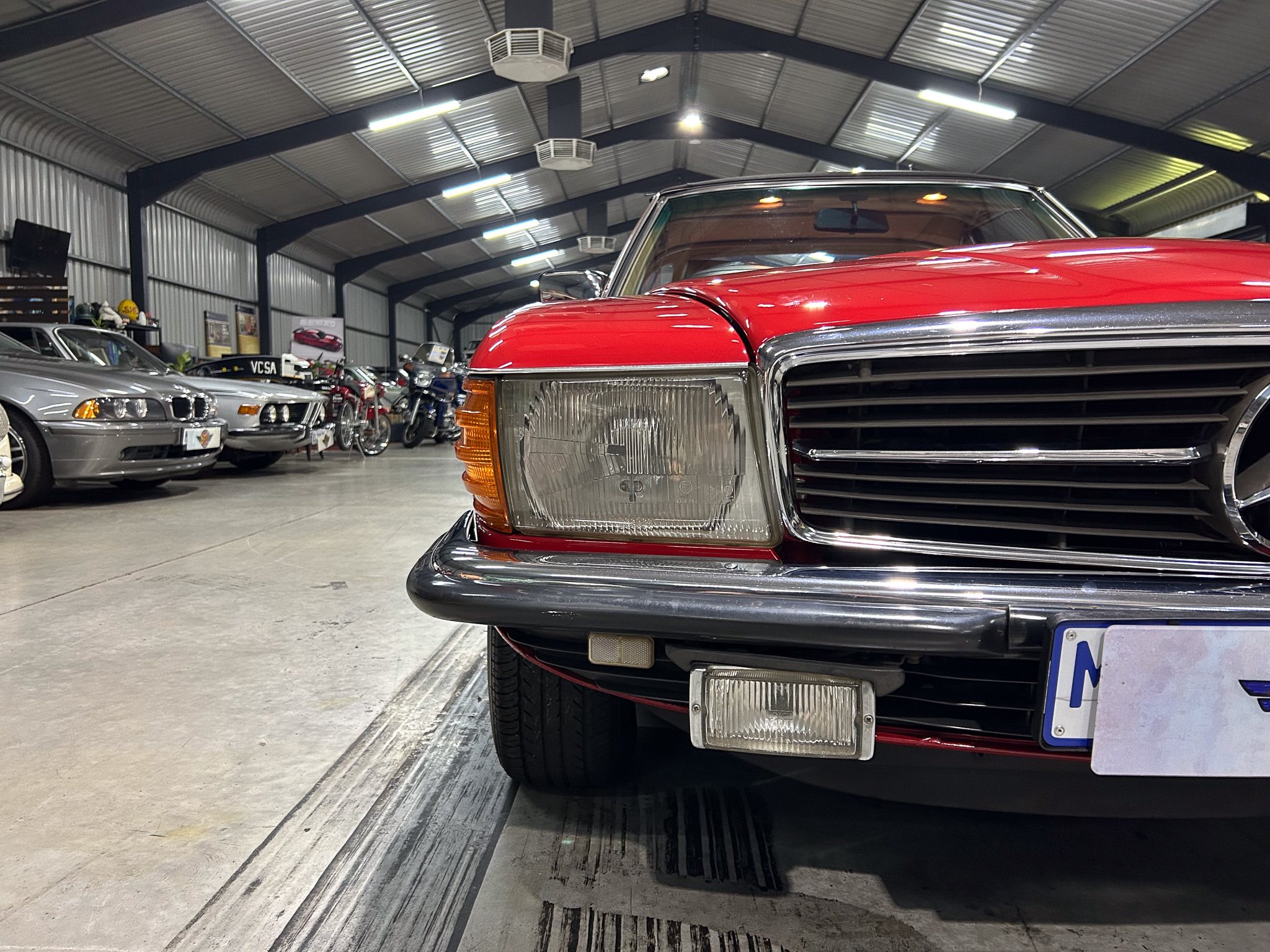 Close-up of the red vintage car’s front end inside a large garage with other classic cars in the background.