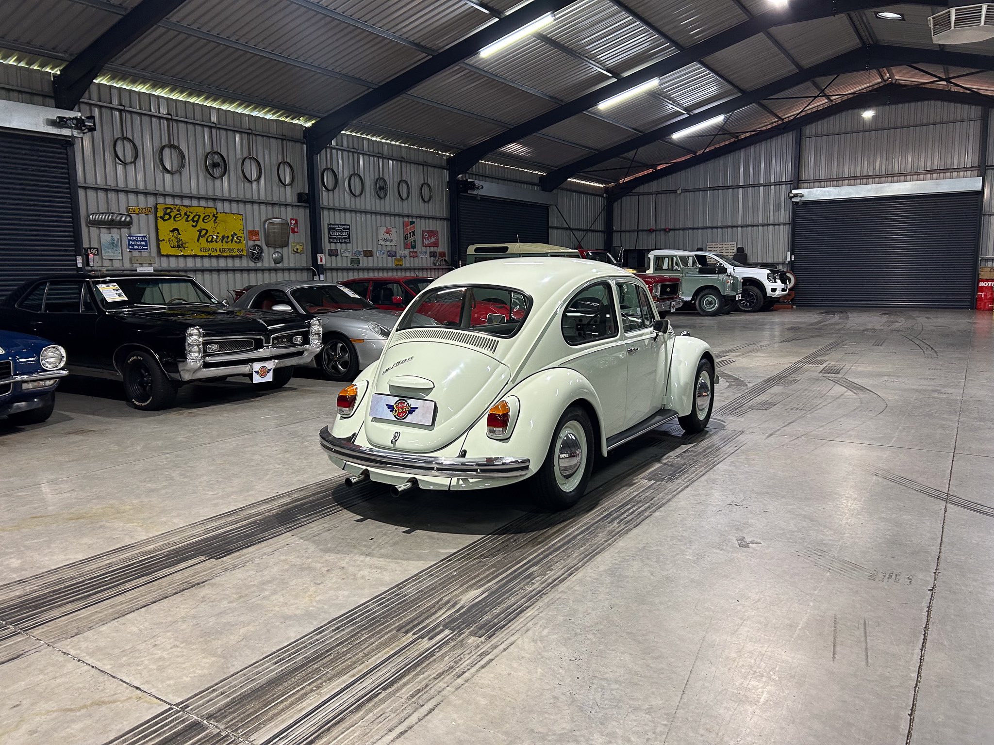 White vintage Volkswagen Beetle in the foreground of a classic car showroom with other cars and metal walls in the background.