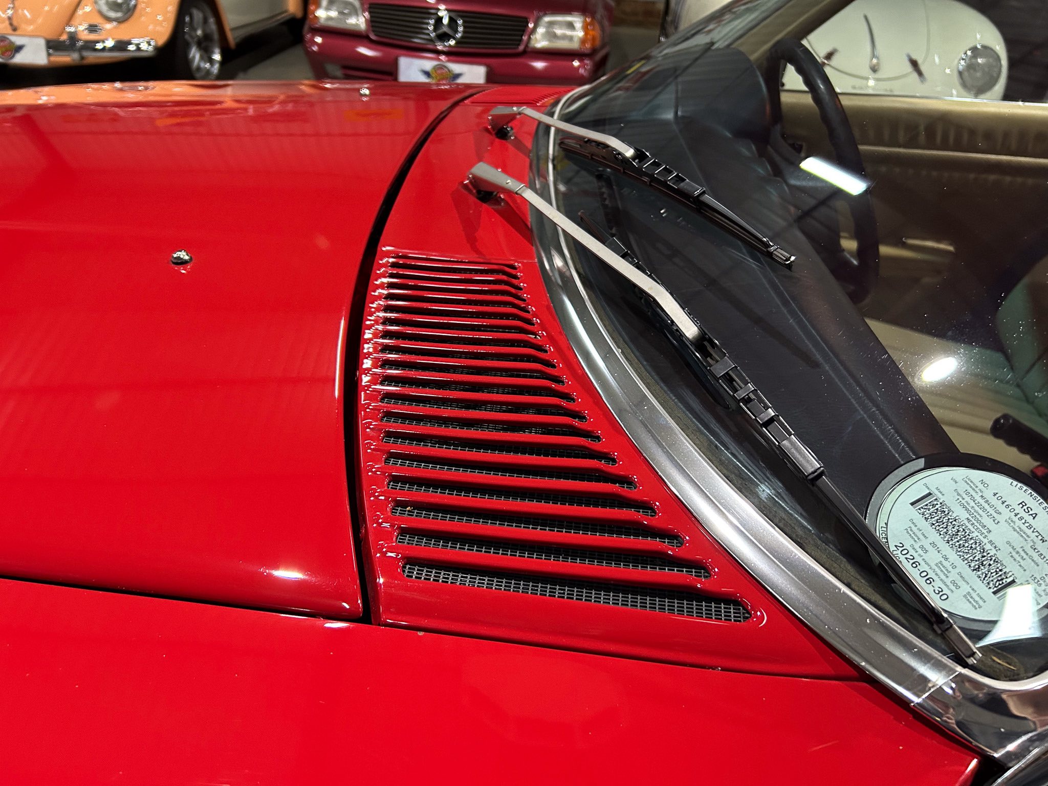 Close-up of a bright red classic car's hood with windshield wipers on the glass; ventilation slats along the side and other vintage cars in the background.
