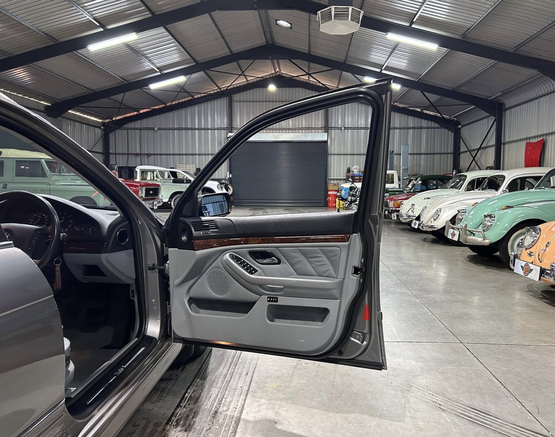 Open car door in a modern SUV with a classic car showroom in the background, lined with vintage vehicles under a metal ceiling.