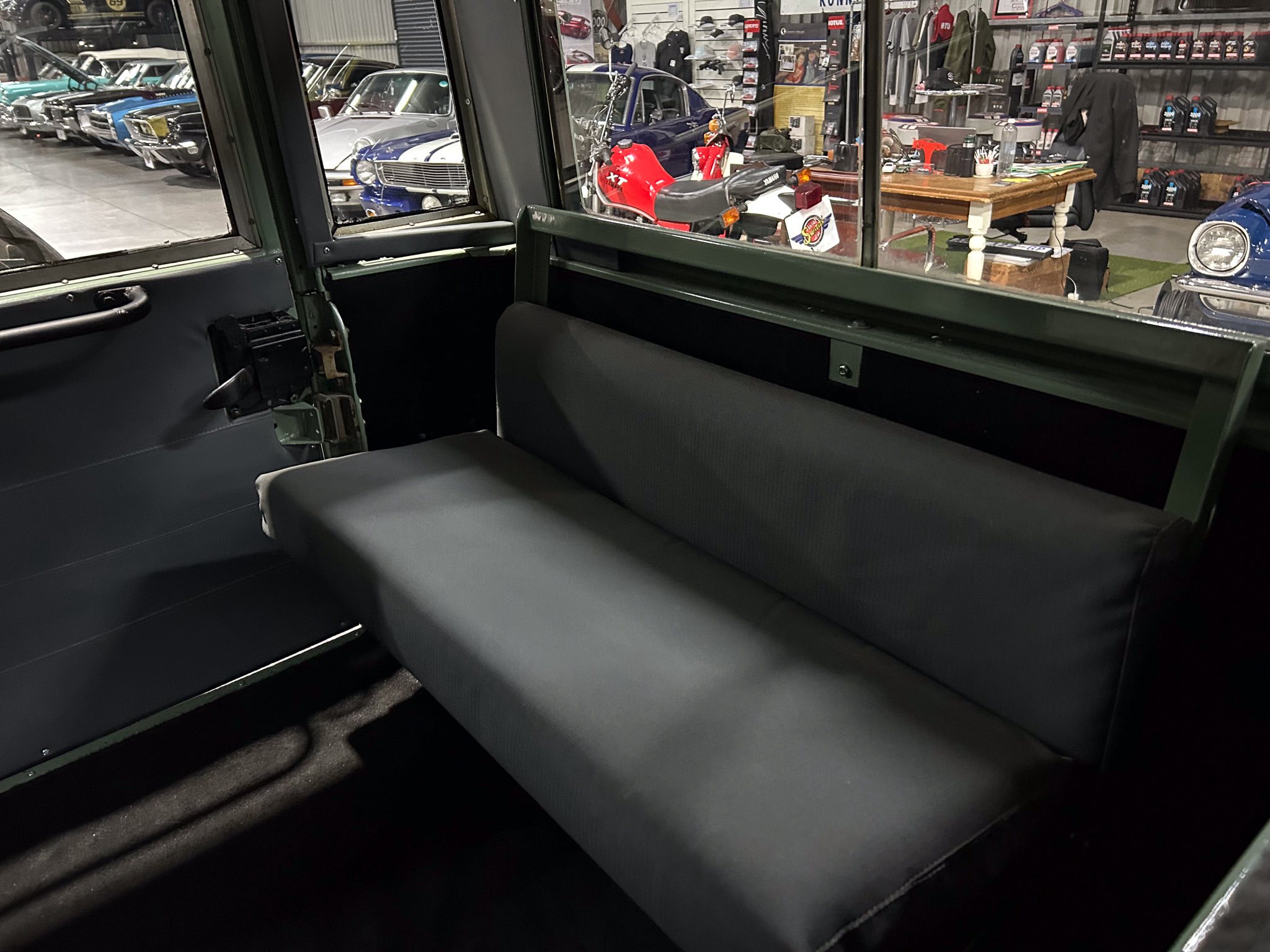 View from the back seat of a vintage car with green metal framing and a black bench seat, showroom of classic cars visible through the window.