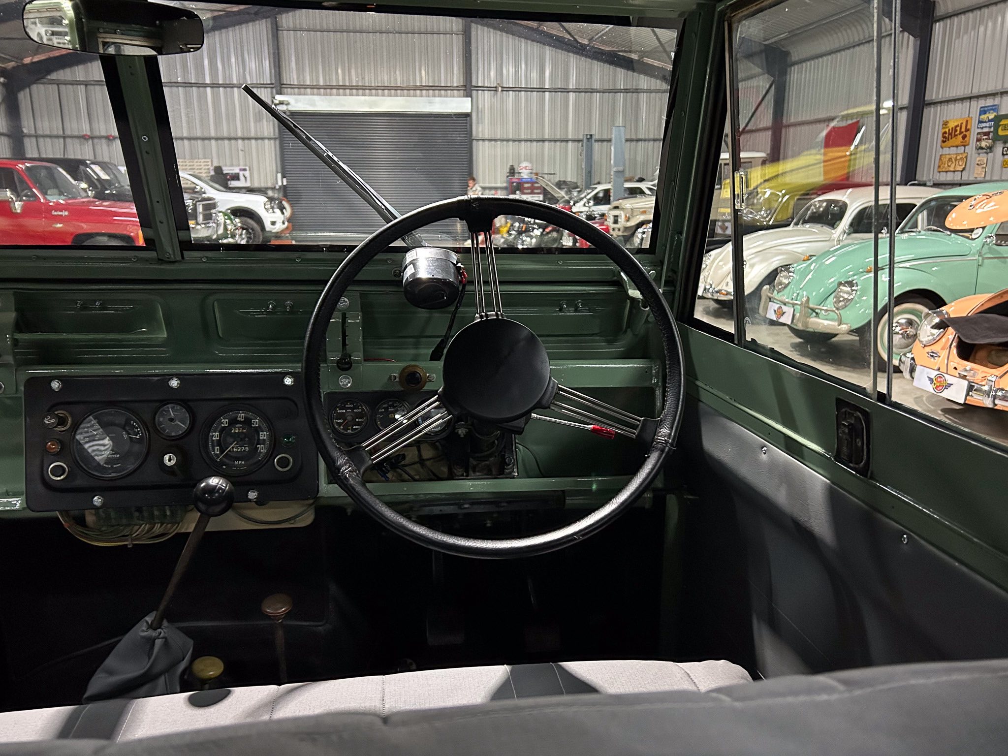 Interior of a vintage green truck showing the steering wheel and gauge cluster, with a showroom of classic cars visible outside the cab window.