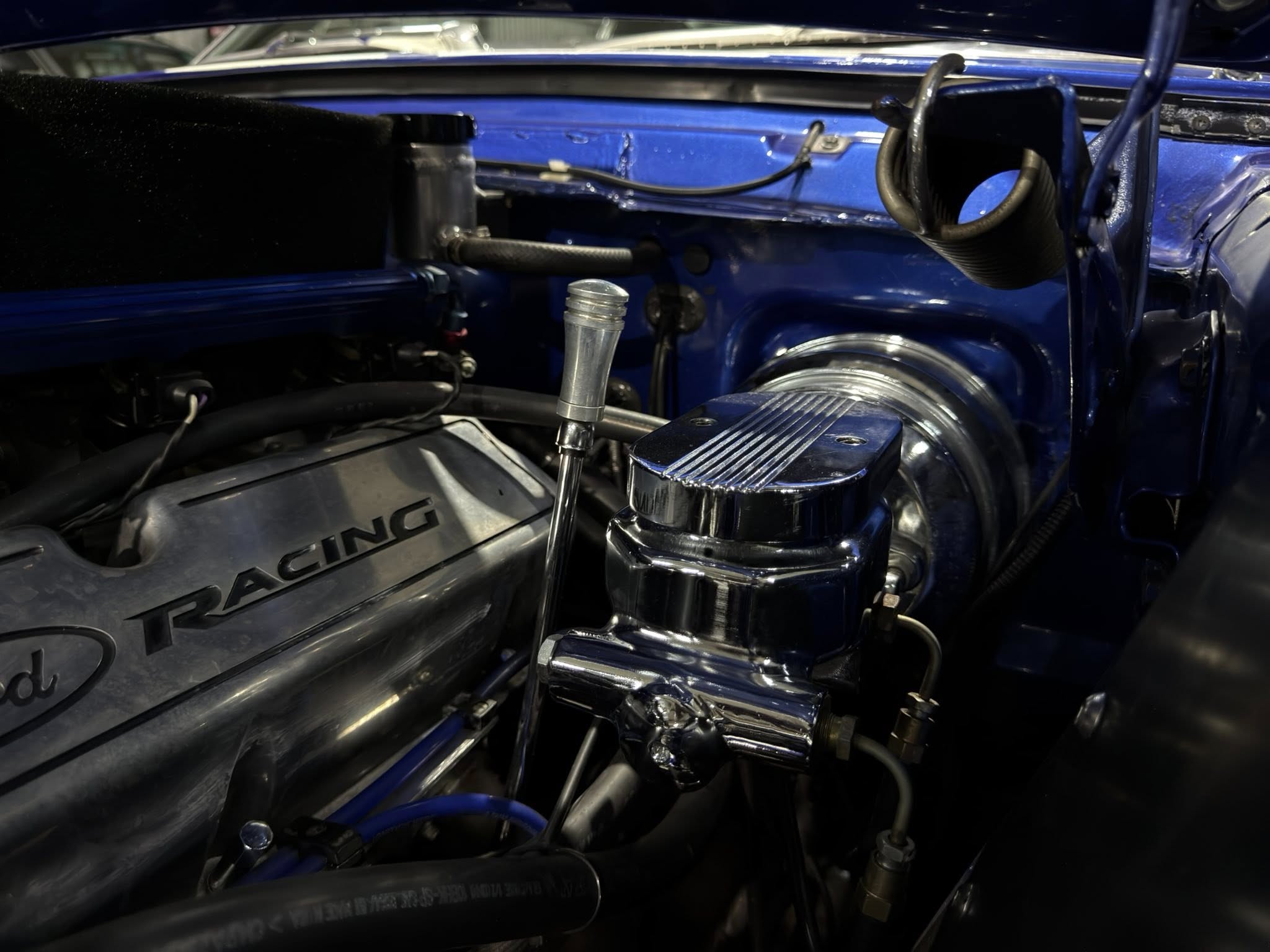 Close-up of a blue car engine bay with hoses, metal fittings, and chrome components visible inside the hood area.