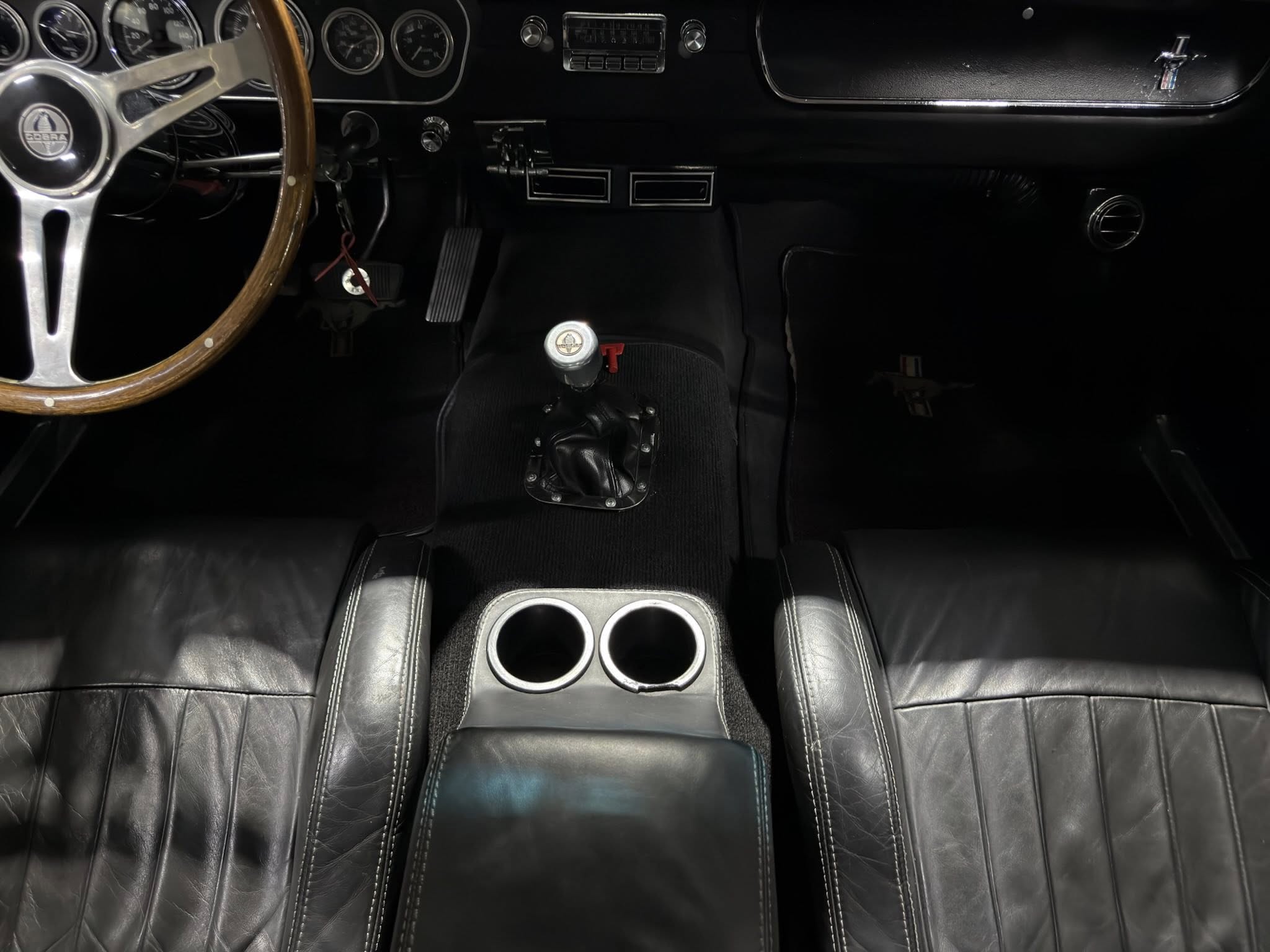 Classic car interior: wood steering wheel, black dashboard, and manual shifter with two-tone center console cups close to front seats.