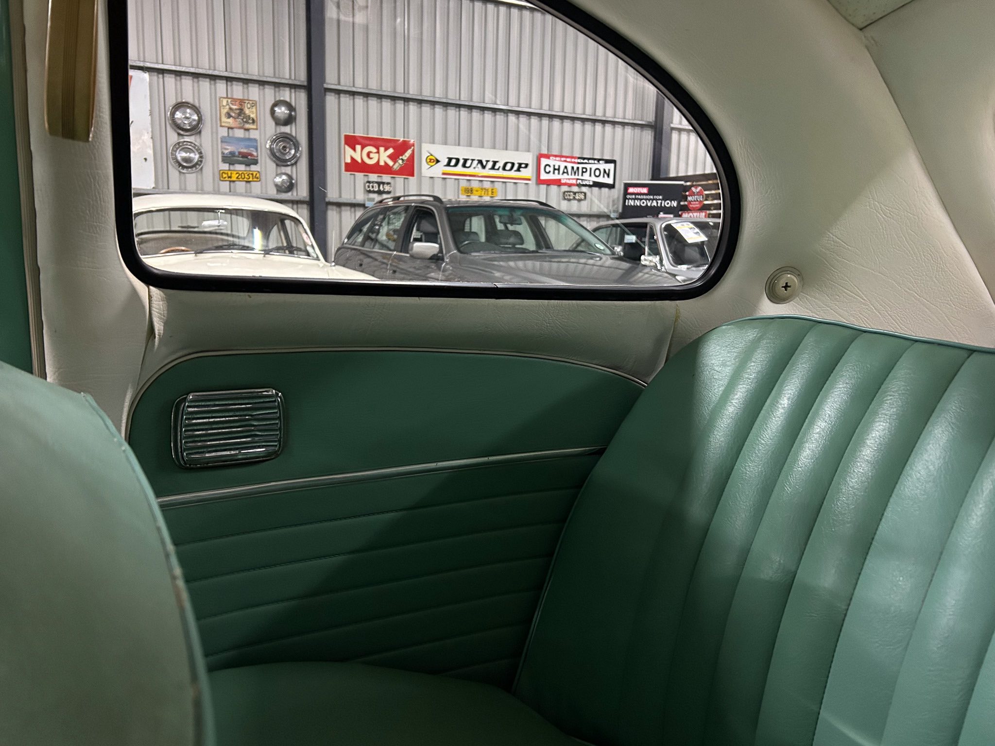 Back seat of a vintage car with green upholstery, viewing a garage wall through the window decorated with auto signs and parked cars outside.