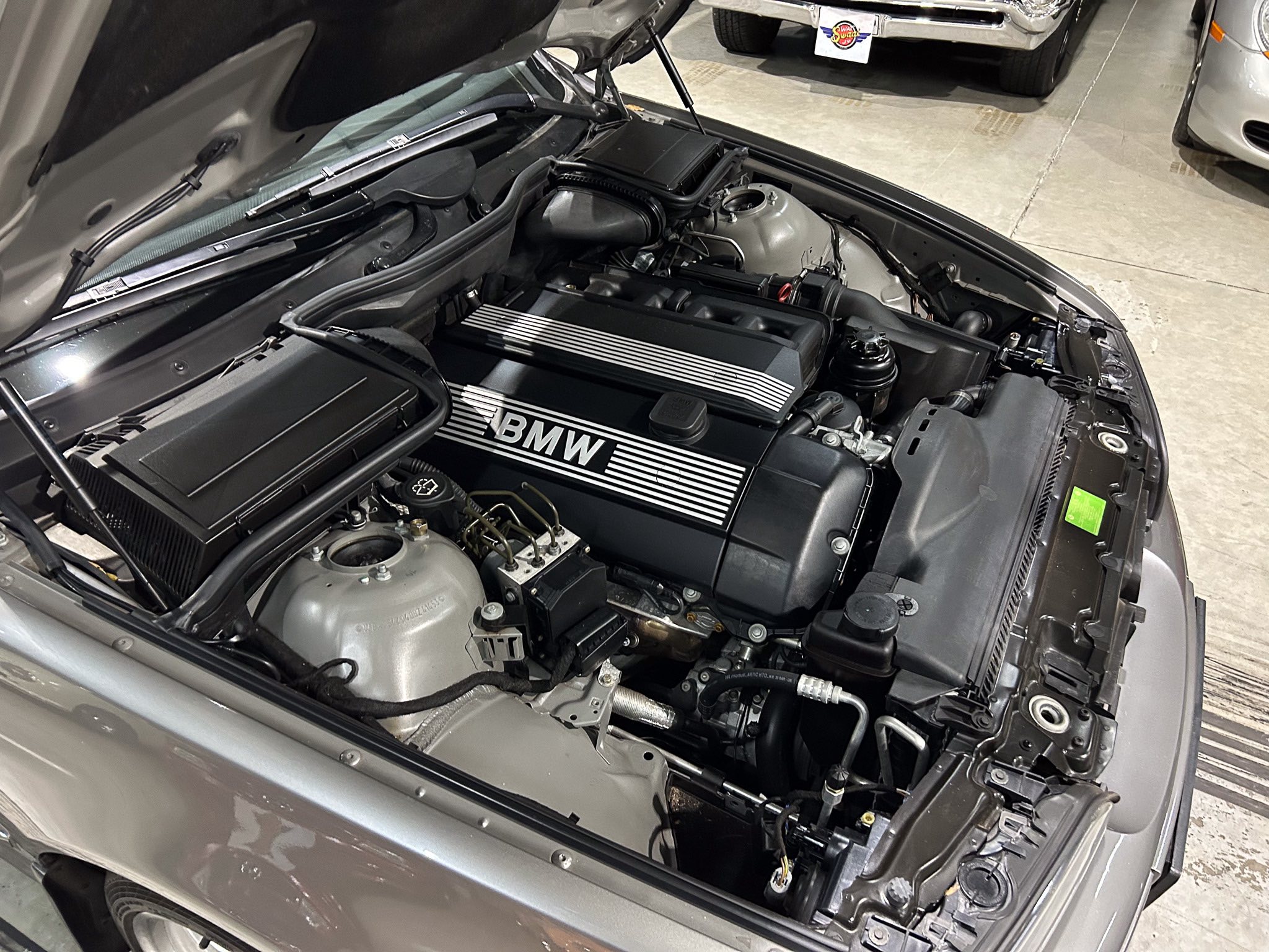 Open hood view of a BMW engine bay, featuring a black intake manifold and a silver striped valve cover with BMW branding visible across the front of the engine.