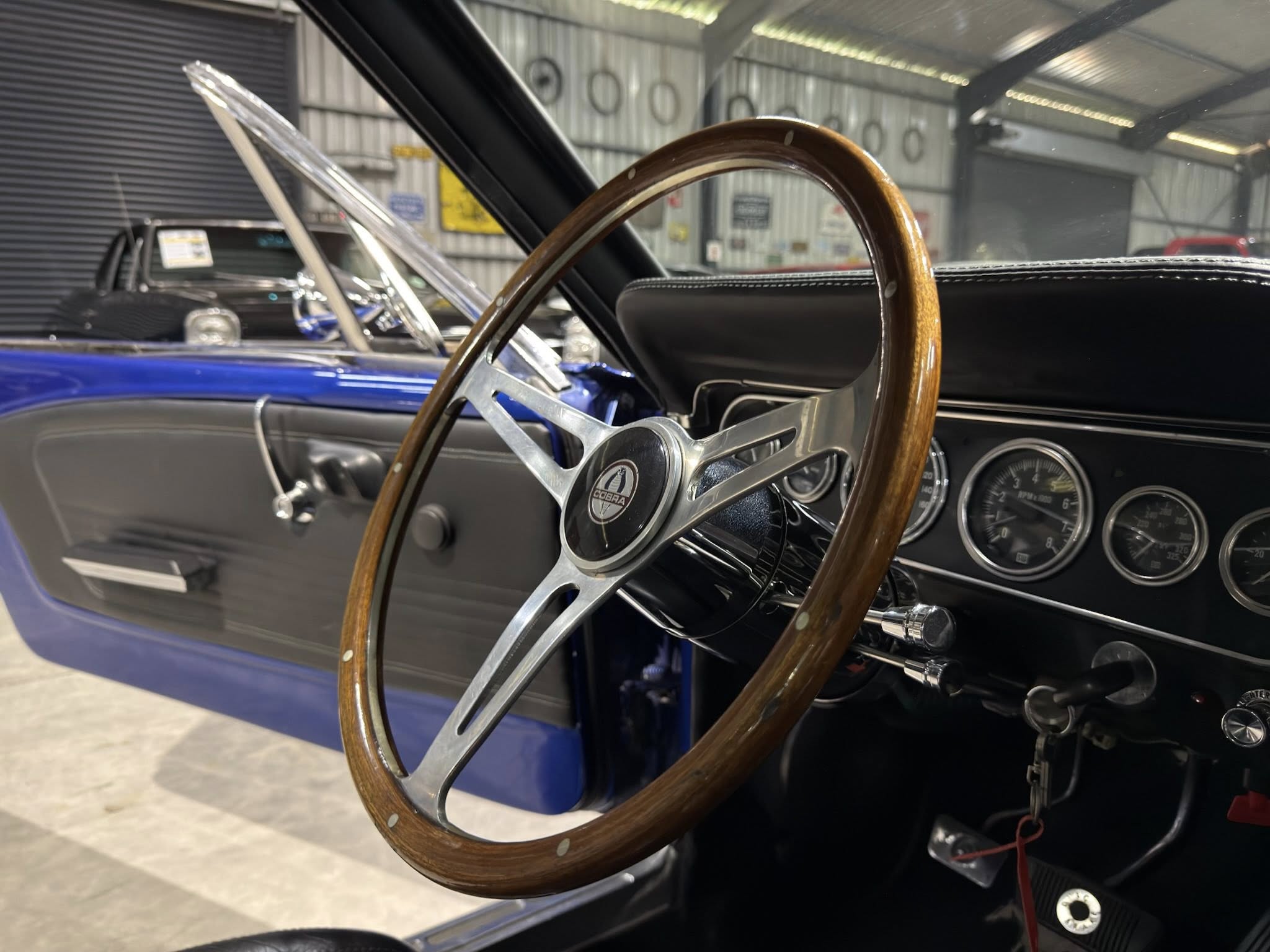 Close-up of a vintage wooden steering wheel inside a blue classic car, showing chrome spokes and gauges in the dashboard inside a garage storage area.