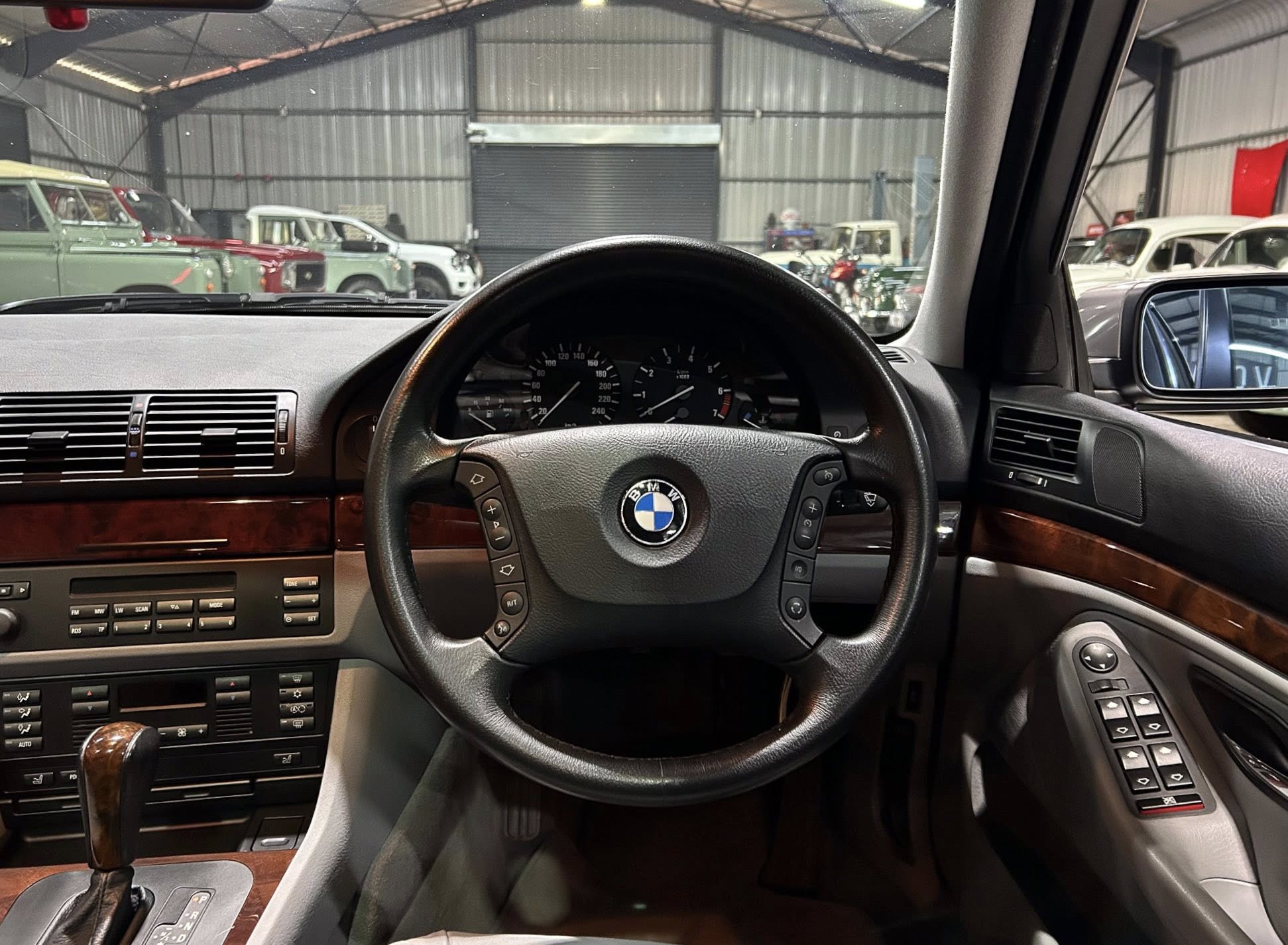 Interior view of a BMW dashboard and steering wheel, with gear shifter and wood trim, inside a garage filled with vintage cars outside the windshield.