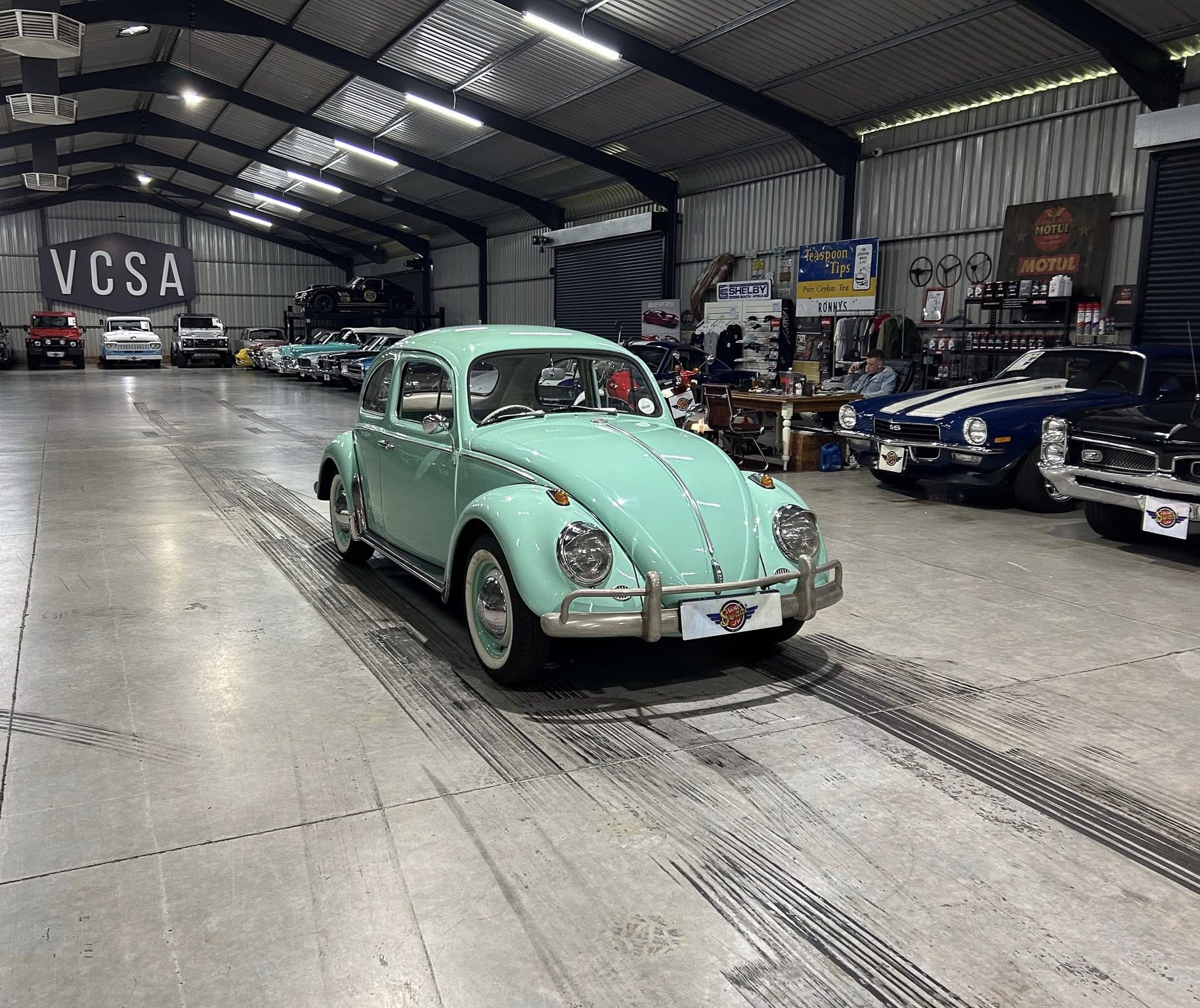 Indoor classic-car showroom with a mint-green Volkswagen Beetle in the foreground and vintage cars lining the back wall.