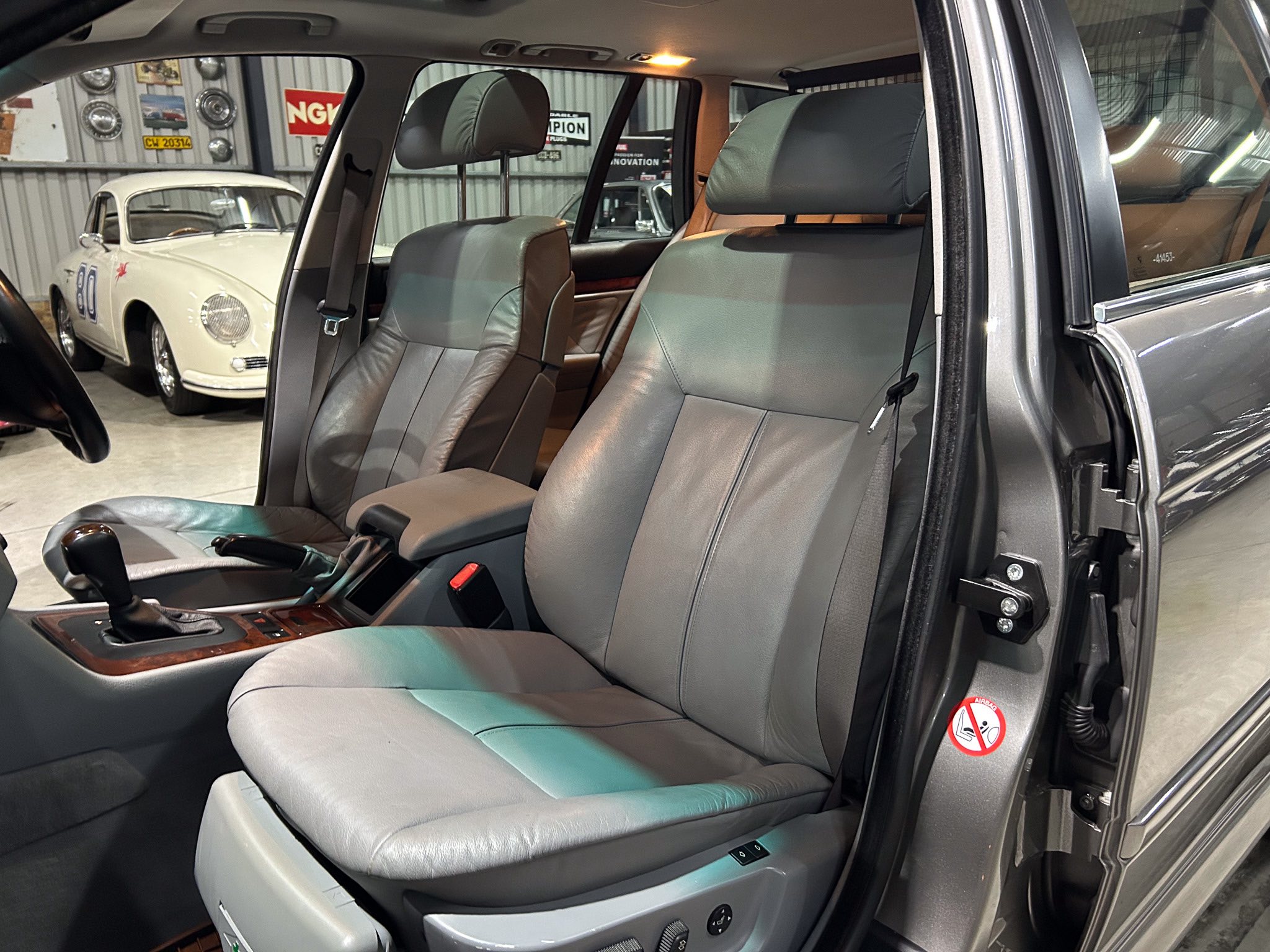 Interior of a gray leather SUV showing front and middle row seats with headrests and center console, open rear door, and a white vintage car visible in a showroom background.