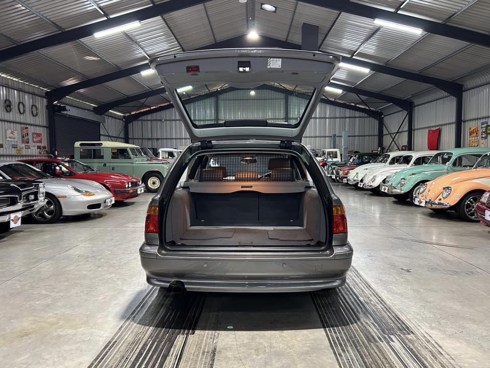 Open hatchback of a silver station wagon in a classic car showroom, trunk raised and interiors visible, surrounded by vintage cars on both sides.