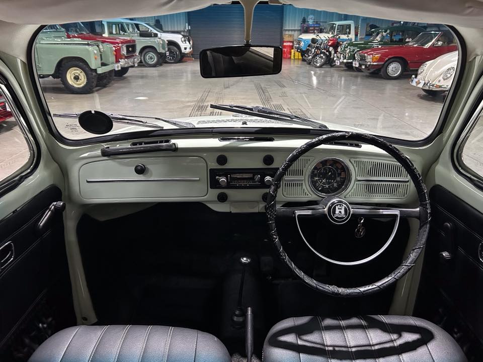 Right-hand-drive vintage car interior with a round speedometer, light-green dashboard, and black seats; showroom filled with classic cars visible through the windshield