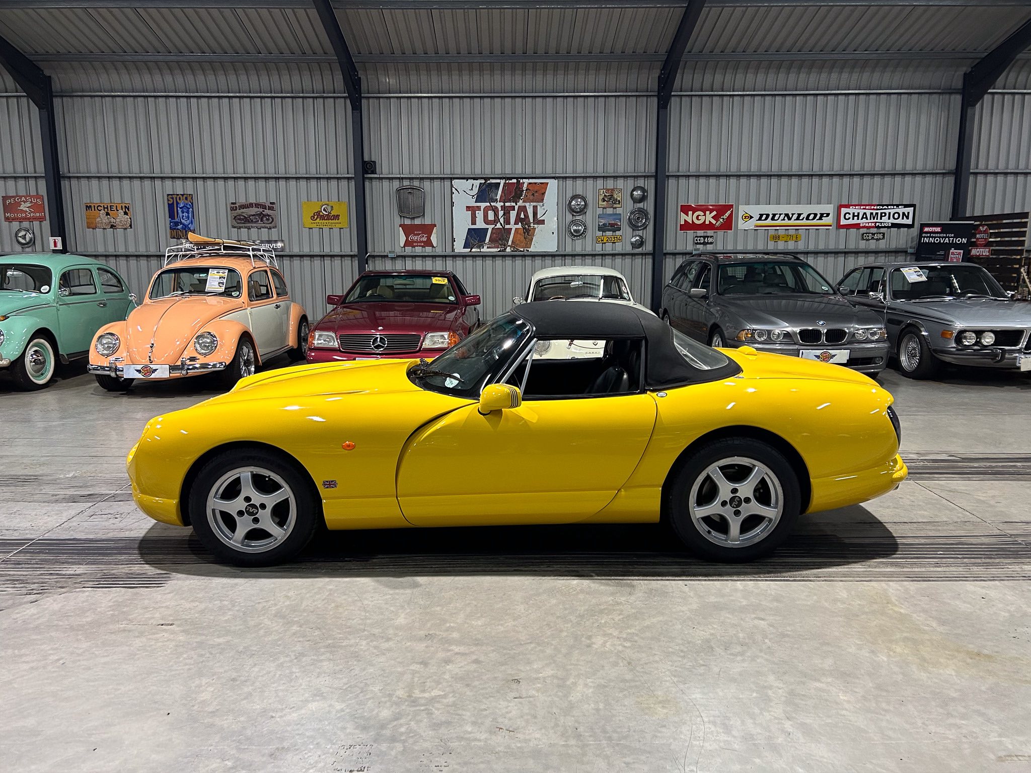 Bright yellow sports car in a showroom with vintage cars and retro wall signs in the background.