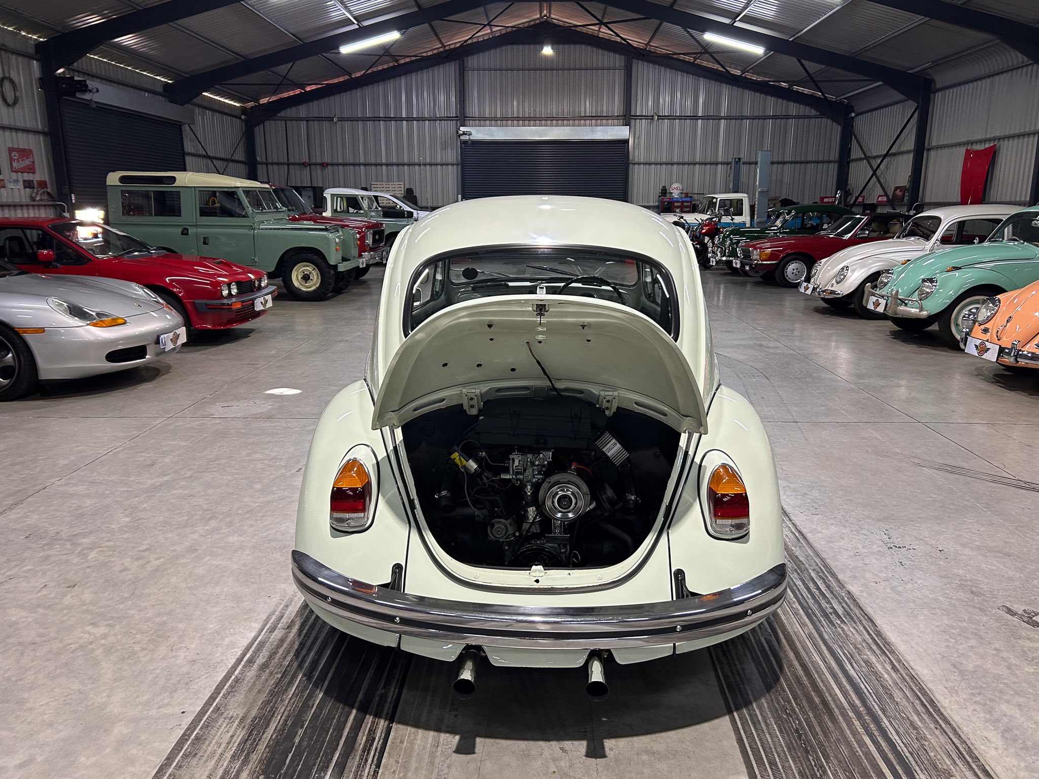 Open engine bay of a pale-green classic car in a spacious warehouse showroom, with rows of vintage vehicles on both sides under metal roofing and bright overhead lights.