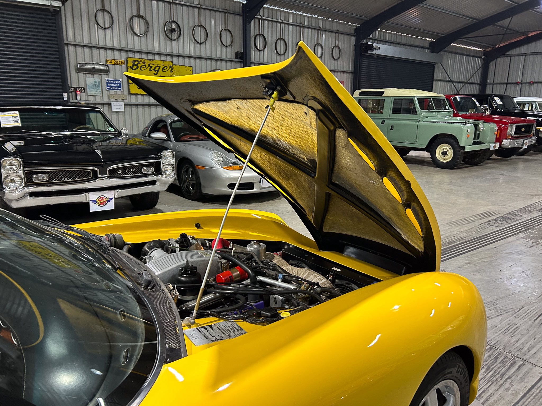 Bright yellow car with its hood raised in a showroom full of classic cars, engine visible in the foreground, black GTO to the left and vintage vehicles to the right behind it.