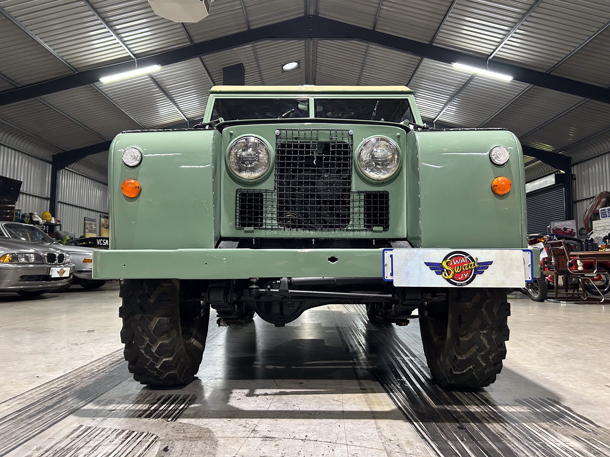 Front view of a green rugged off-road vehicle inside a workshop, showing large tires, round headlights, and a protective grille.