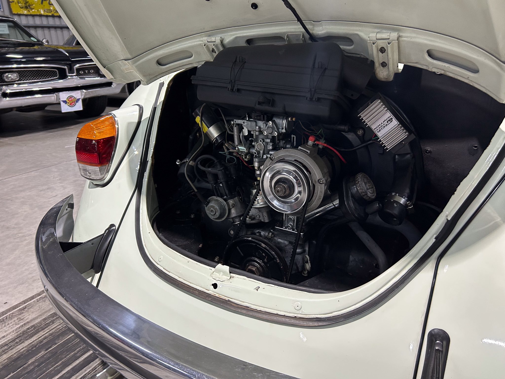 Open rear hatch of a light-colored vintage car, exposing the engine and belts with another classic car visible in the background.