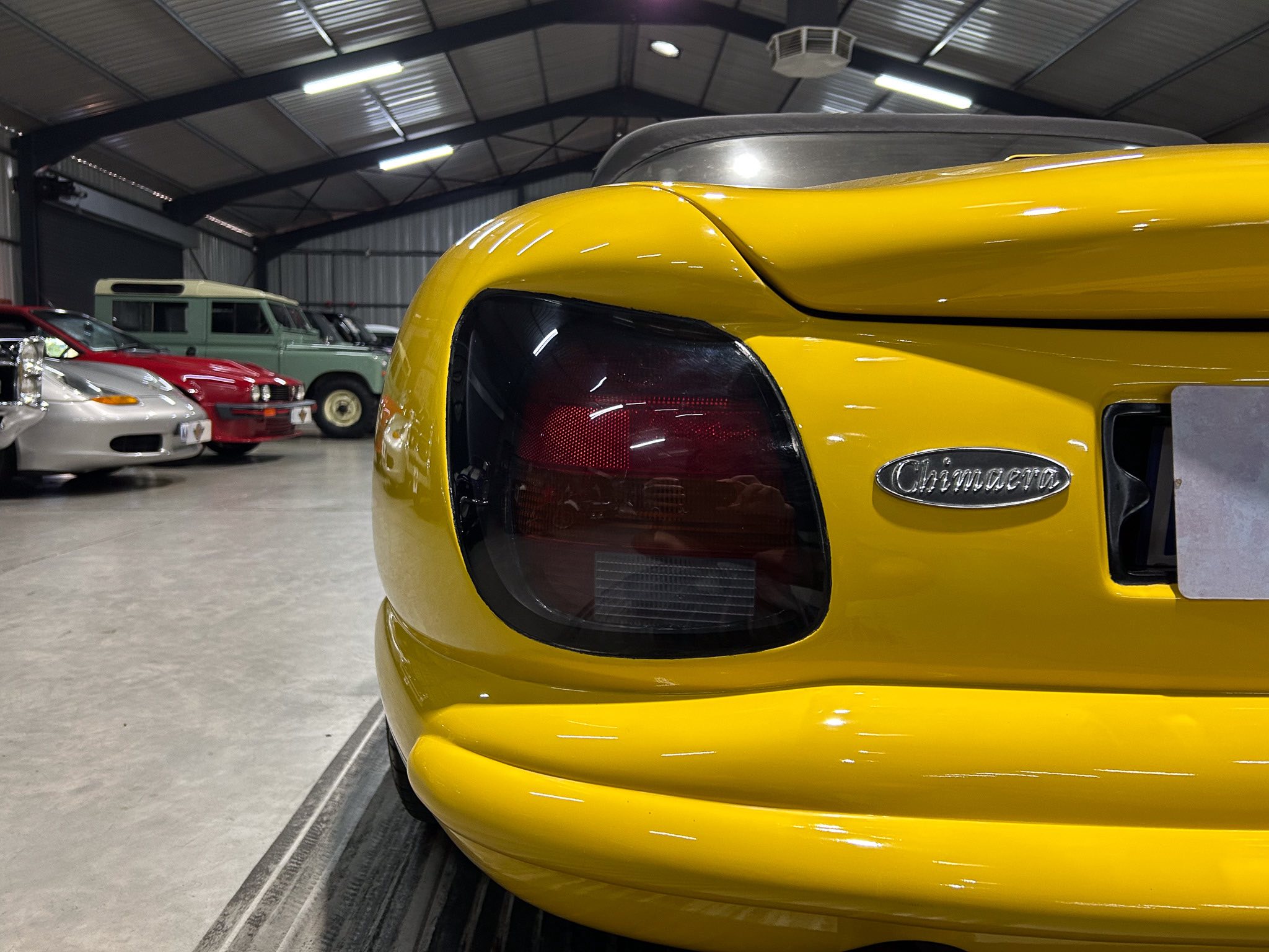 Close-up of a bright yellow car's rear corner with a red taillight and chrome badge in a classic car showroom or garage. In the background, other vintage vehicles are visible under a high, metal-roofed ceiling.