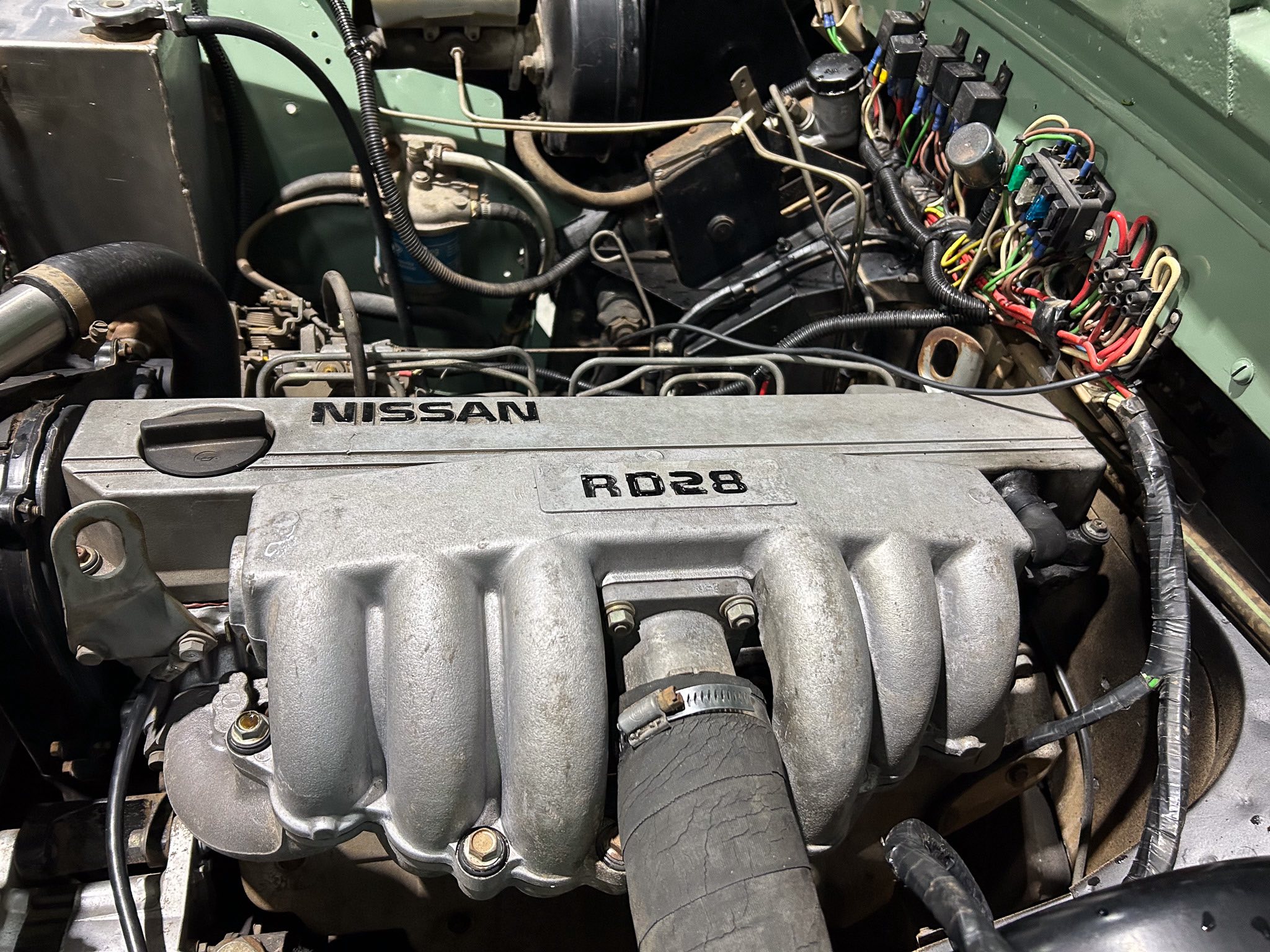 Close-up of a Nissan RD28 engine bay with a tangle of wires, hoses, and components visible in a green engine bay.