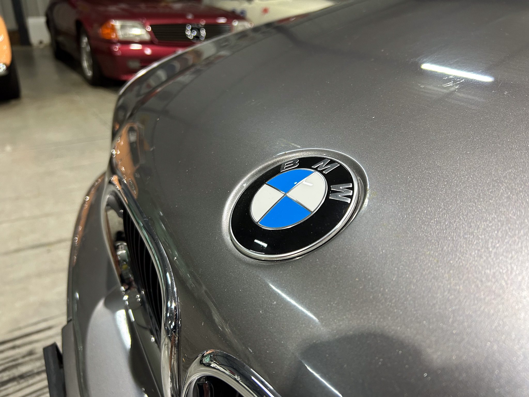 Close-up of the BMW emblem on a gray car hood with a blurred red car in the background