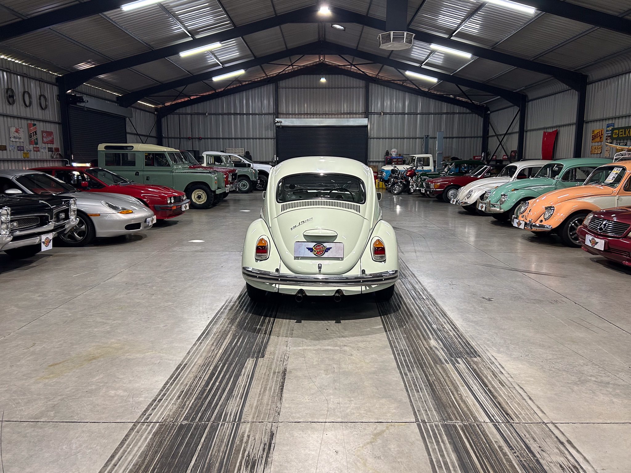Indoor car showroom with vintage cars lined along both sides; white VW Beetle centered in the foreground.